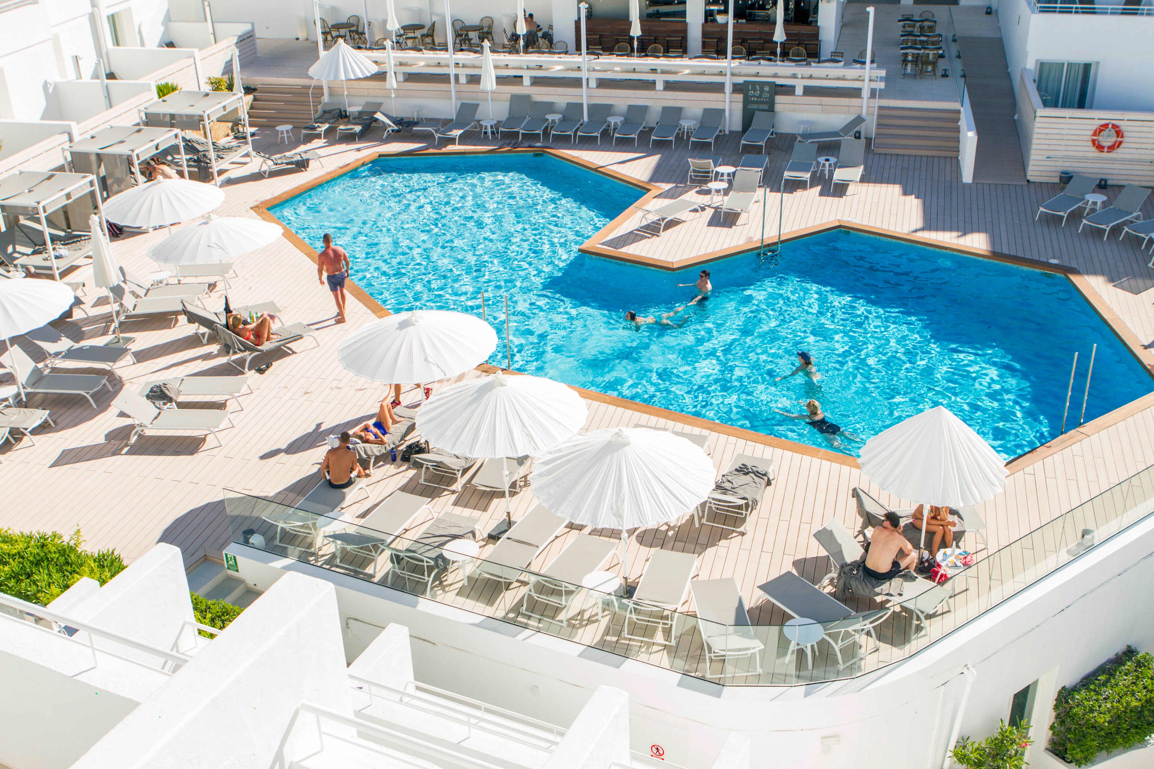 An elevated view shows a bright resort swimming pool surrounded by sun loungers and white umbrellas. Several people relax on deck chairs while others swim and play in the clear blue water. The pool deck and surrounding architecture are light-colored, enhancing the sunny, vacation atmosphere. The scene suggests a warm day at a modern hotel or holiday resort.