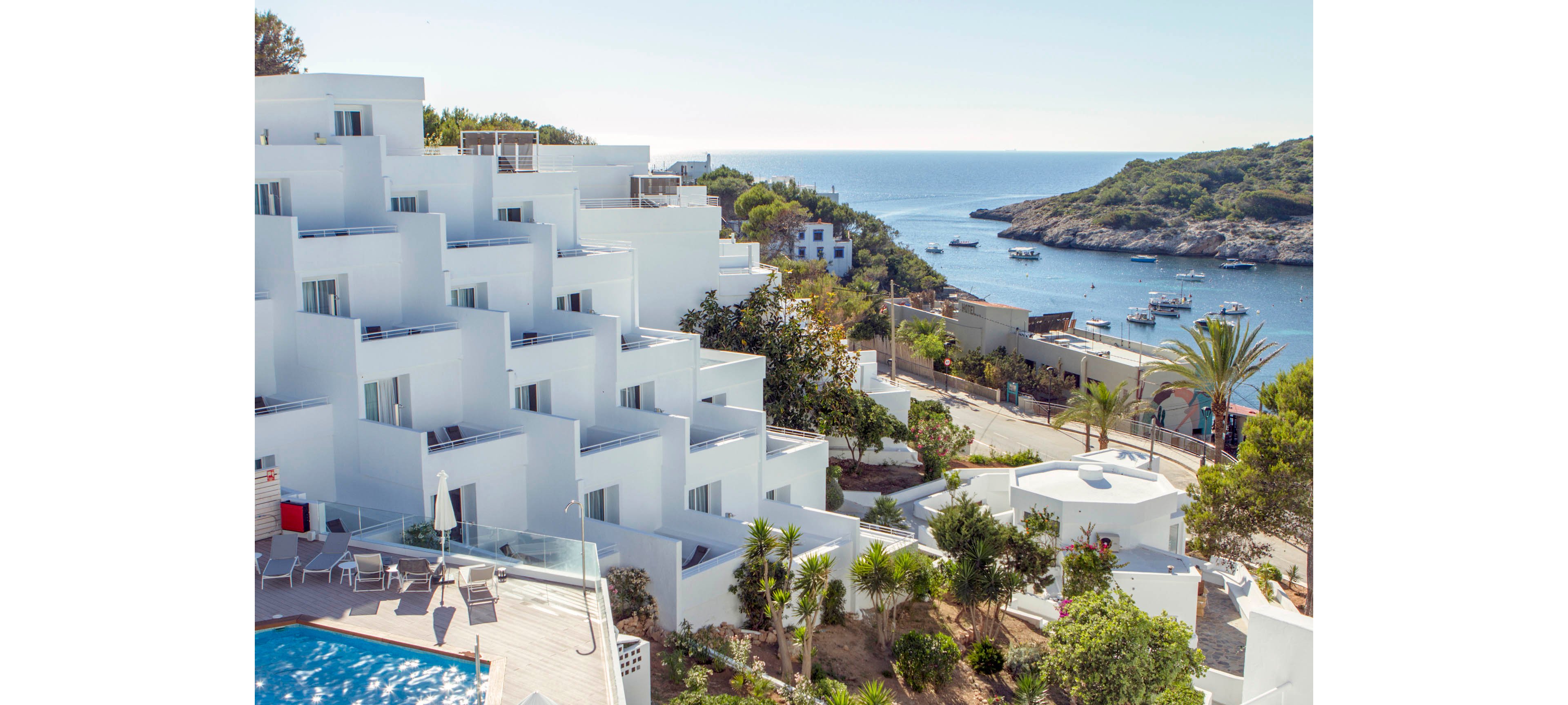 A modern white hotel complex with stepped balconies overlooks a calm coastal bay. The building features clean geometric lines and multiple terraces facing the sea. In the foreground, a swimming pool area with lounge chairs is surrounded by palm trees and greenery. Beyond the hotel, a small harbor with boats and rocky shoreline stretches into the distance under a clear sky.