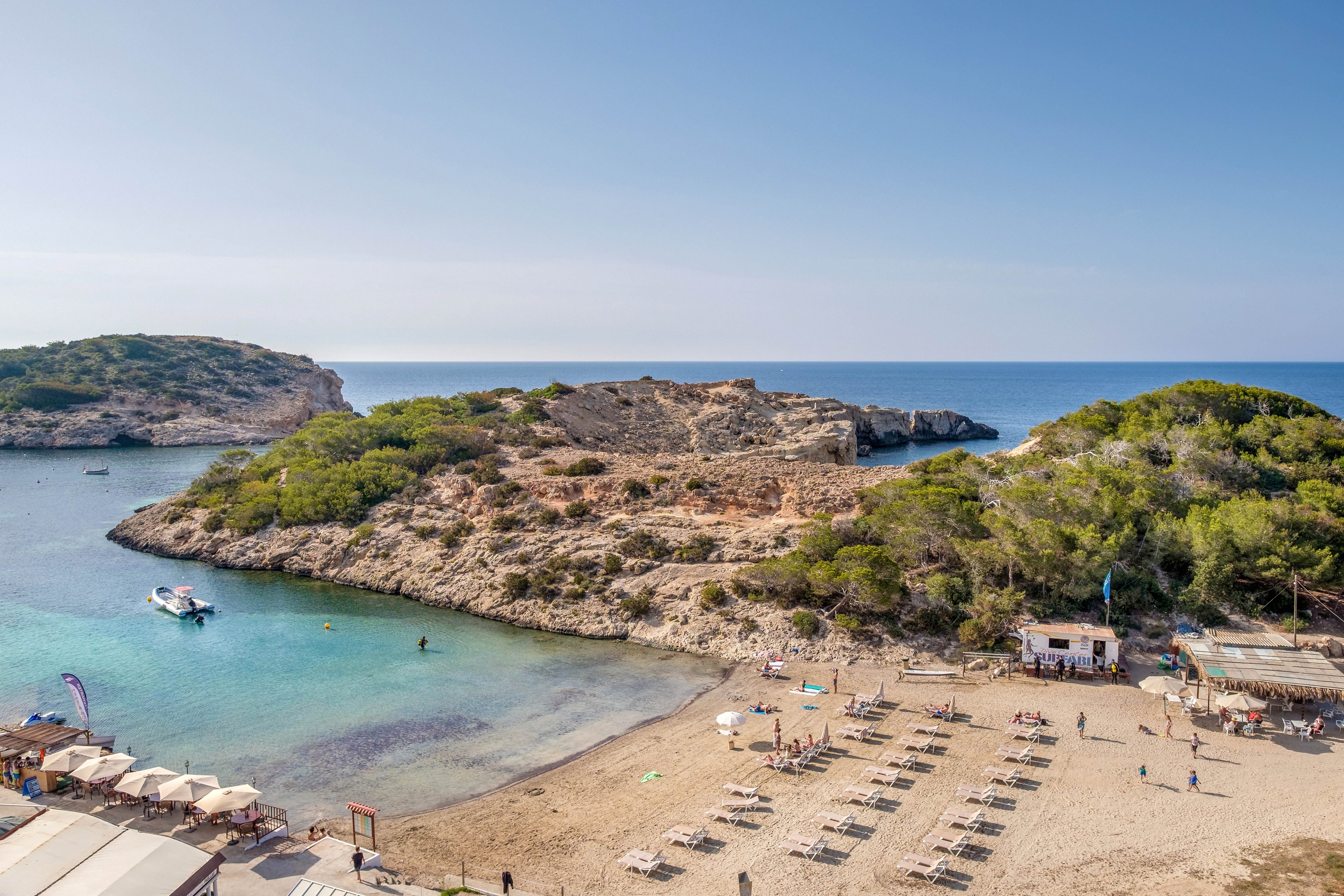 A tranquil sandy beach curves around a clear turquoise cove bordered by rocky cliffs and lush green trees. Neatly arranged sun loungers and umbrellas line the shoreline, with a few people scattered along the sand and near the water. A small boat floats in the calm sea close to the rocky outcrop, adding to the relaxed coastal atmosphere. Beachfront buildings and a beach bar sit at the edge of the sand, enhancing the holiday resort feel.