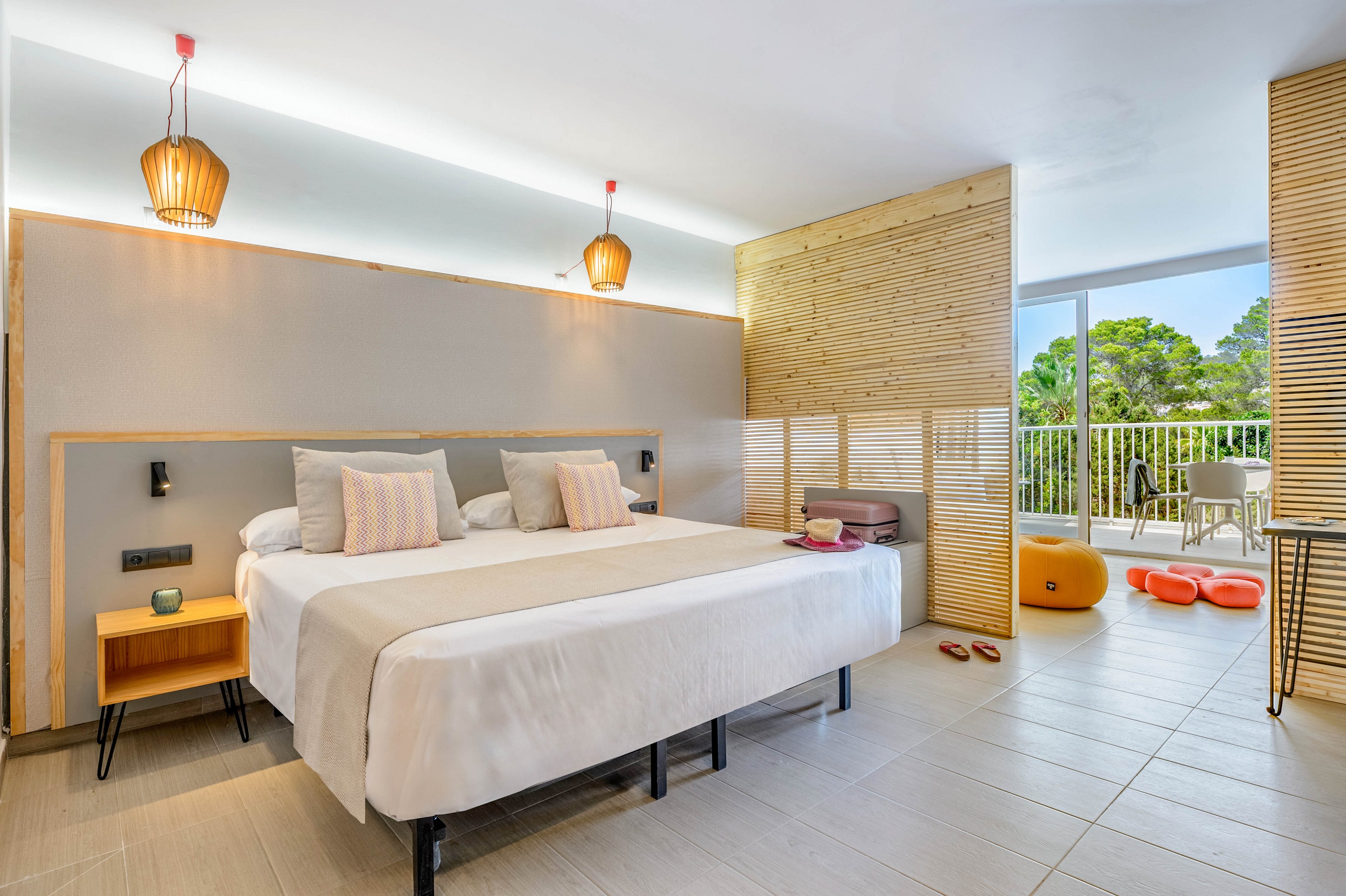 Contemporary hotel room featuring a large double bed with neutral bedding and striped accent pillows. The space is decorated with warm wooden panels, minimalist bedside tables, and hanging pendant lights. A sliding glass door opens to a balcony with outdoor seating and views of green trees. Colorful suitcases and pool floats add a relaxed vacation atmosphere to the clean, sunlit interior.