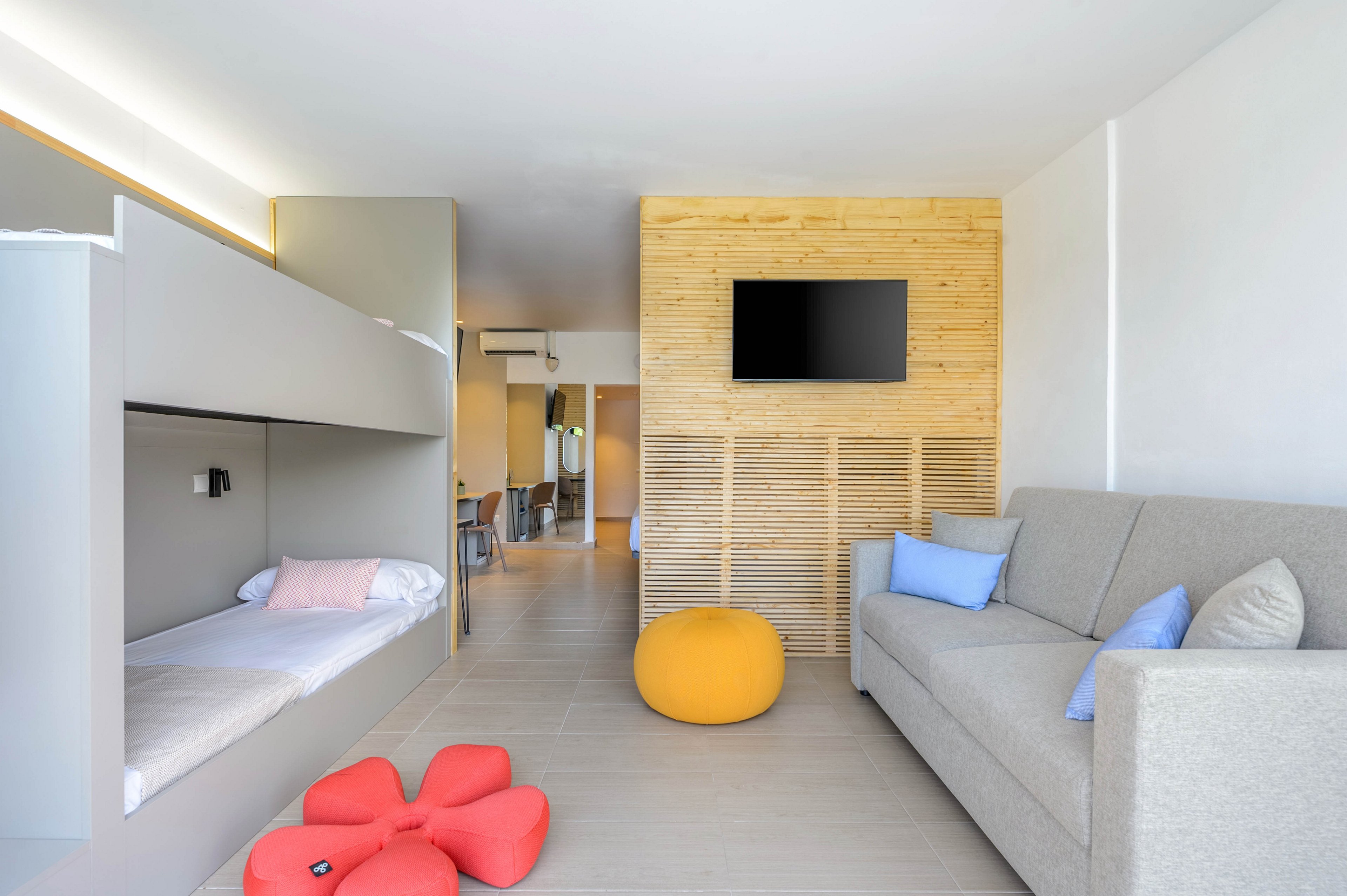 Bright contemporary hotel room featuring built-in bunk beds and a light gray sofa. A wooden accent wall with a flat-screen TV adds warmth to the minimalist interior. Colorful cushions and poufs in yellow, blue, and red provide playful contrast against neutral tones. The space appears clean, spacious, and designed for families or groups.