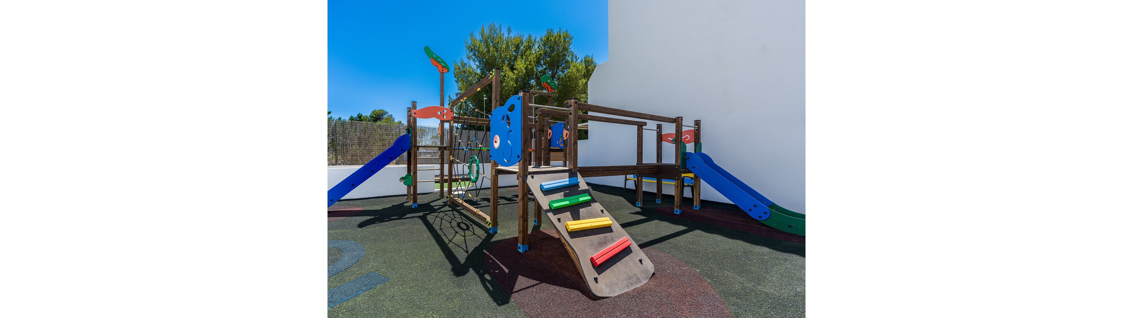 An empty outdoor playground features a wooden climbing structure under a clear blue sky. The play set includes a slide, climbing ramp with colorful steps, and a rope net. Bright red, blue, yellow, and green elements stand out against the dark rubber safety flooring. Trees and a white building wall frame the background, suggesting a sunny, warm day.