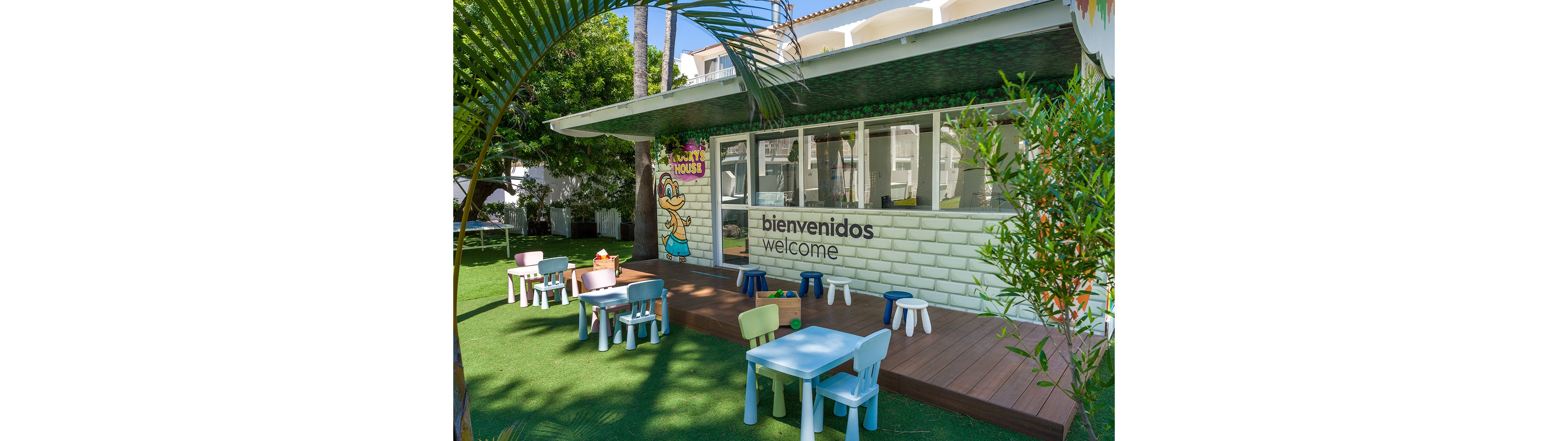 An outdoor children's play area features small colorful tables and stools arranged on artificial grass. A white brick hut with large windows stands behind the seating, with the words "bienvenidos" and "welcome" clearly visible on the wall. A cartoon character and playful decor suggest a fun, family-friendly environment. Lush green plants and trees surround the space, creating a bright and inviting atmosphere for kids.