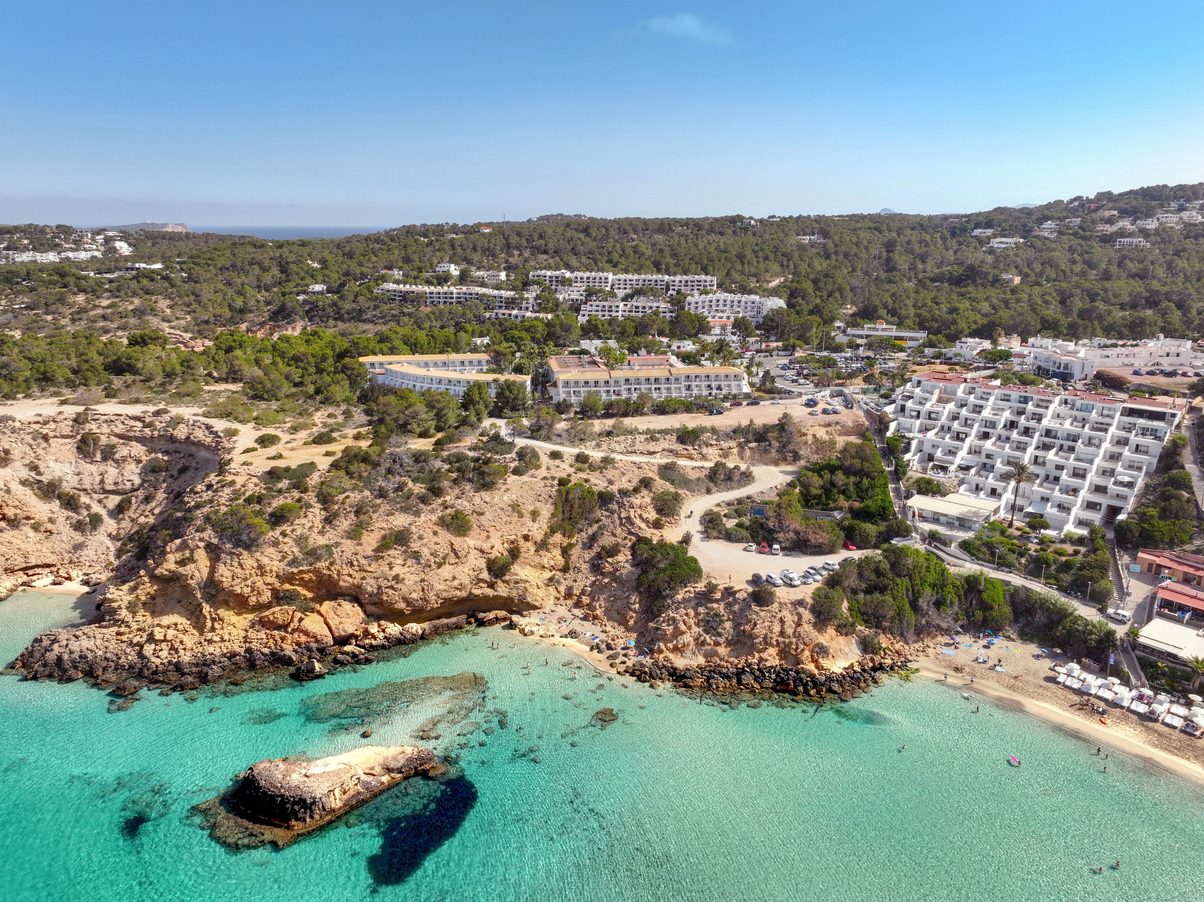 A turquoise bay with clear water and rocky cliffs is seen from above, with a small rock formation near the shore. White resort buildings and hotels line the hillside overlooking the beach, surrounded by dense green forest. Sun loungers and umbrellas are visible along the sandy shoreline, indicating a popular vacation destination. The bright daylight and clear blue sky create a warm, inviting summer atmosphere.