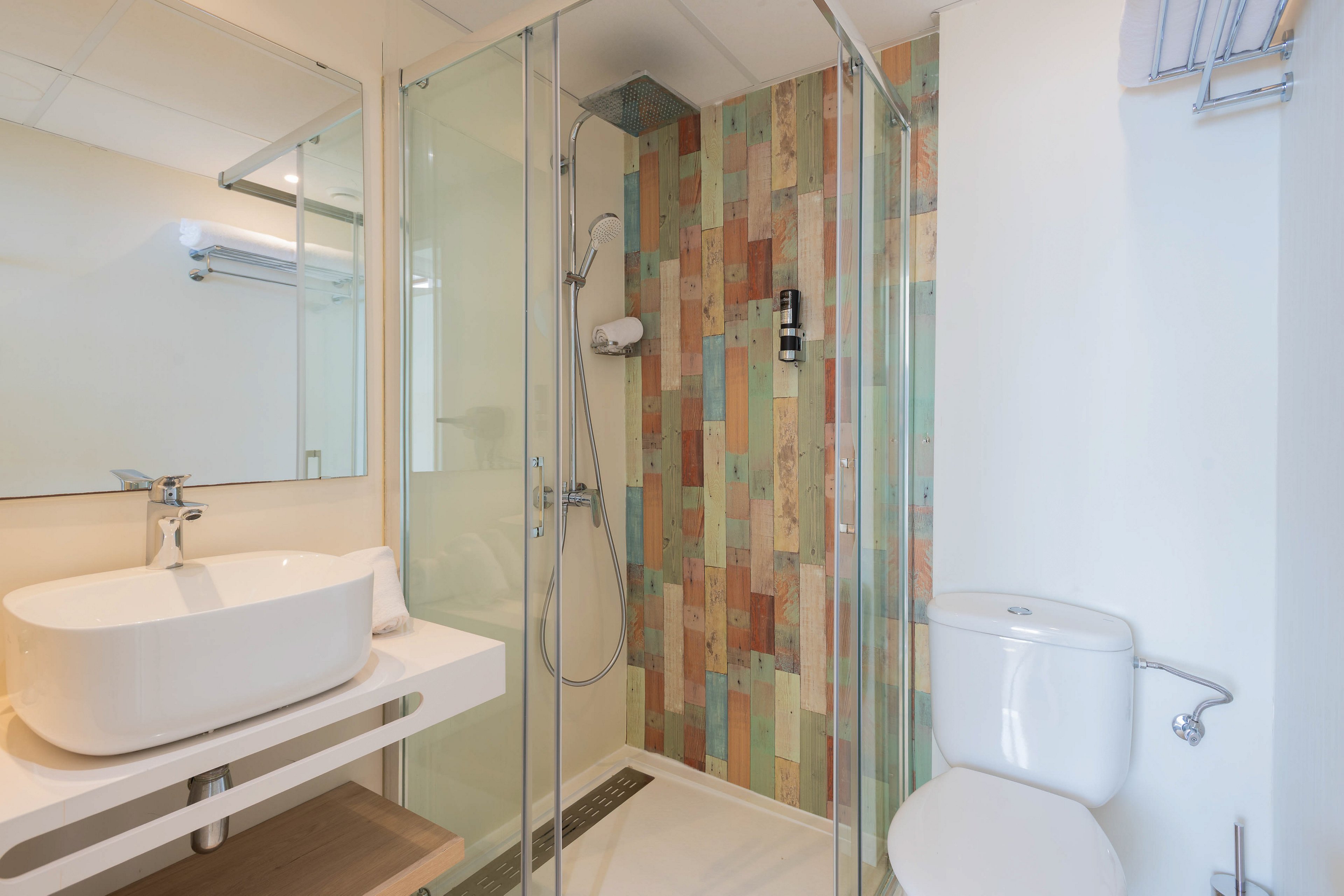 A contemporary bathroom featuring a glass-enclosed shower with vibrant, multicolored tiles. The space includes a sleek white sink on a minimalist vanity and a white toilet. The room is well-lit with natural and artificial lighting, creating a clean and inviting atmosphere.