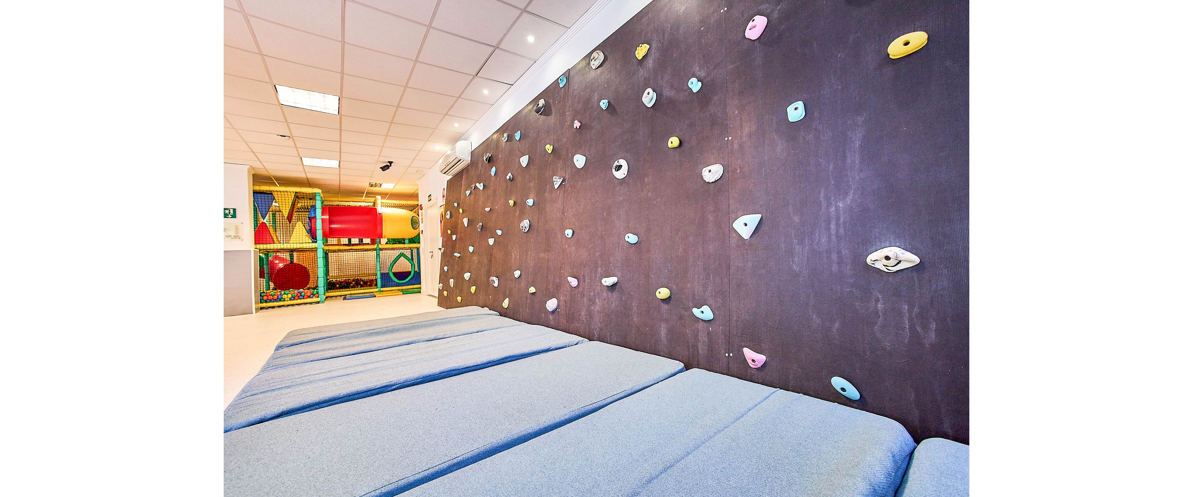 An indoor climbing wall with colorful holds stretches along one side of a bright playroom. Thick padded mats cover the floor beneath the wall for safety. In the background, a soft play structure with tunnels and shapes adds to the children’s activity space. The room is well lit with a grid of ceiling lights, creating a clean and inviting environment.