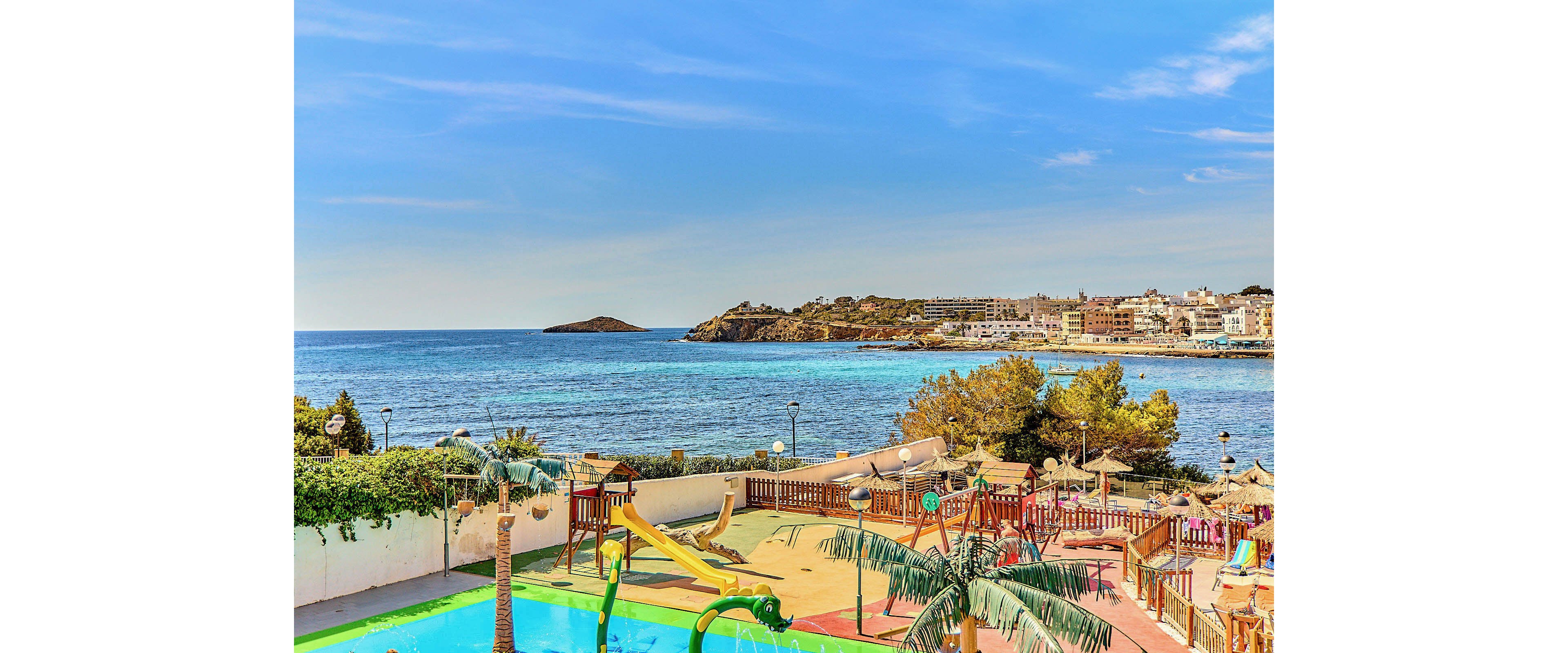 A colorful resort pool area overlooks a calm blue sea. The foreground features a swimming pool, water slide, and a children’s playground with palm-like decorations. Sun loungers and parasols are arranged along the terrace, suggesting a relaxed holiday atmosphere. In the distance, a coastal town and small island sit under a bright, clear sky.