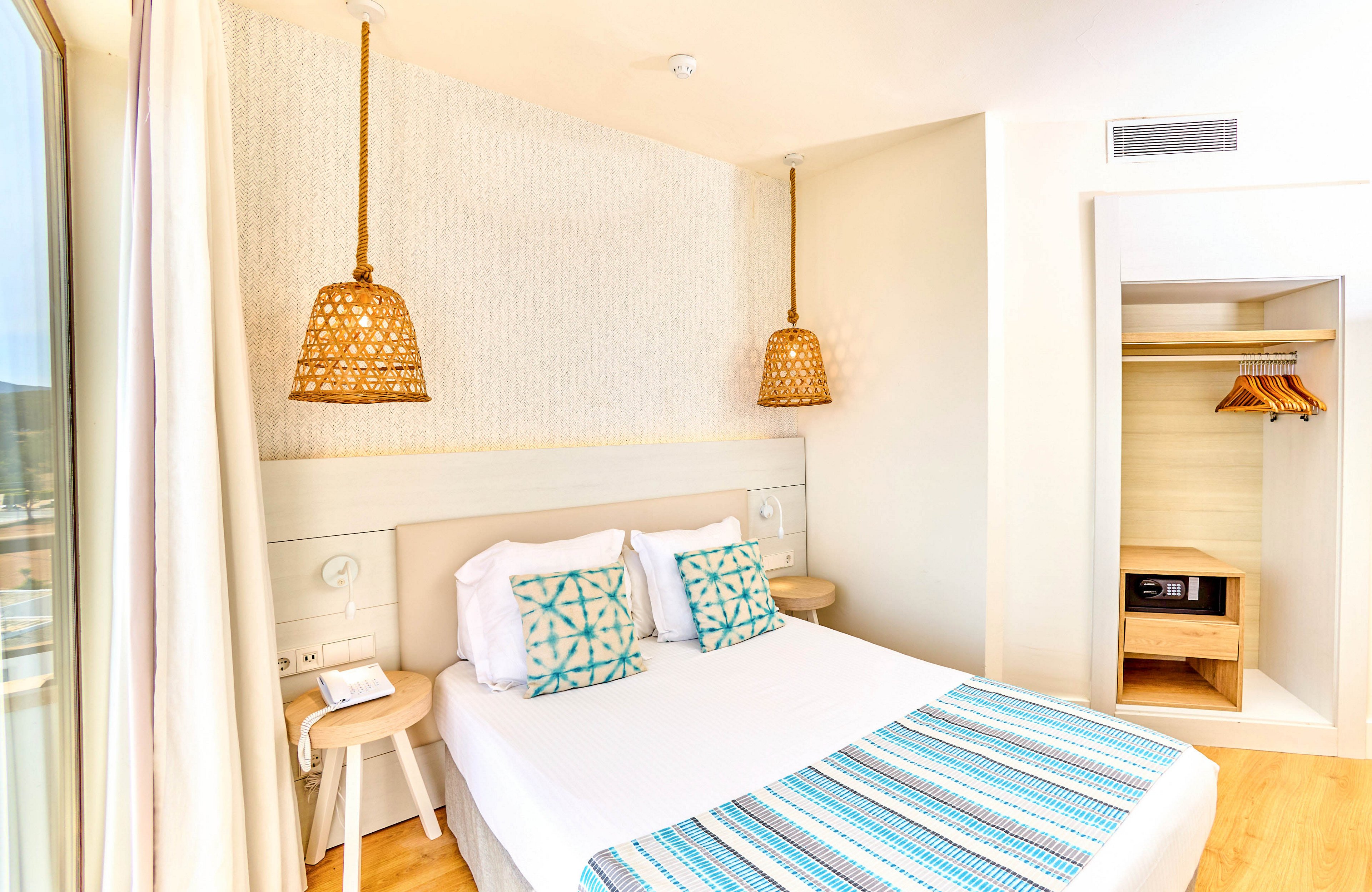 A sunlit hotel bedroom features a neatly made double bed with patterned pillows and a matching striped throw. Woven pendant lights hang above minimalist bedside tables, adding a warm, natural touch. An open closet with wooden hangers and a small safe or storage unit is visible in the corner. Large windows suggest access to a balcony and fill the room with bright daylight, emphasizing the clean, contemporary decor.