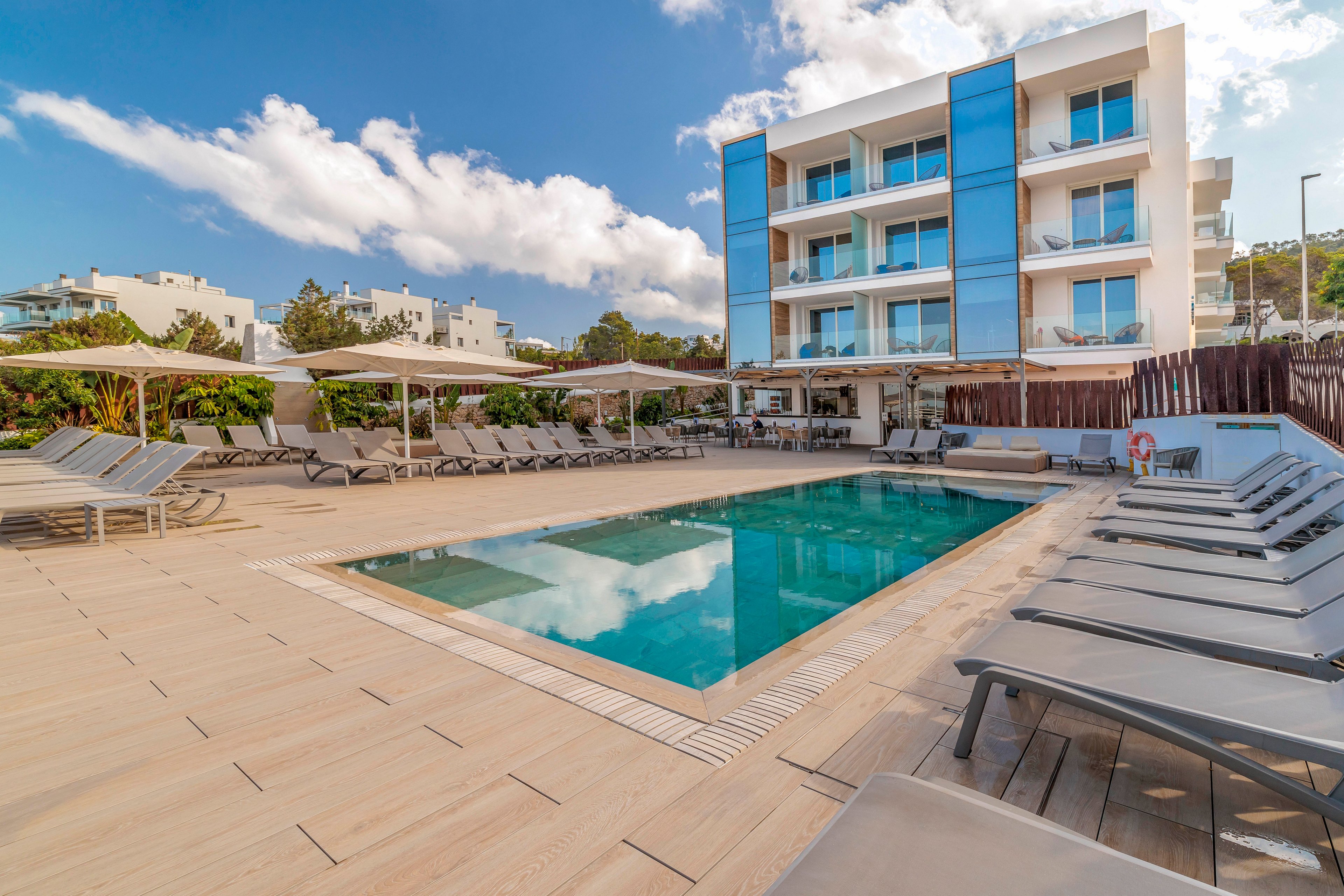 A contemporary hotel building overlooks a rectangular outdoor swimming pool surrounded by sun loungers. The scene is set on a spacious wooden deck with parasols providing shaded seating areas. The architecture features clean lines, glass balconies, and bright blue panels that reflect the clear sky. Nearby residential buildings and lush greenery frame the relaxed vacation atmosphere.