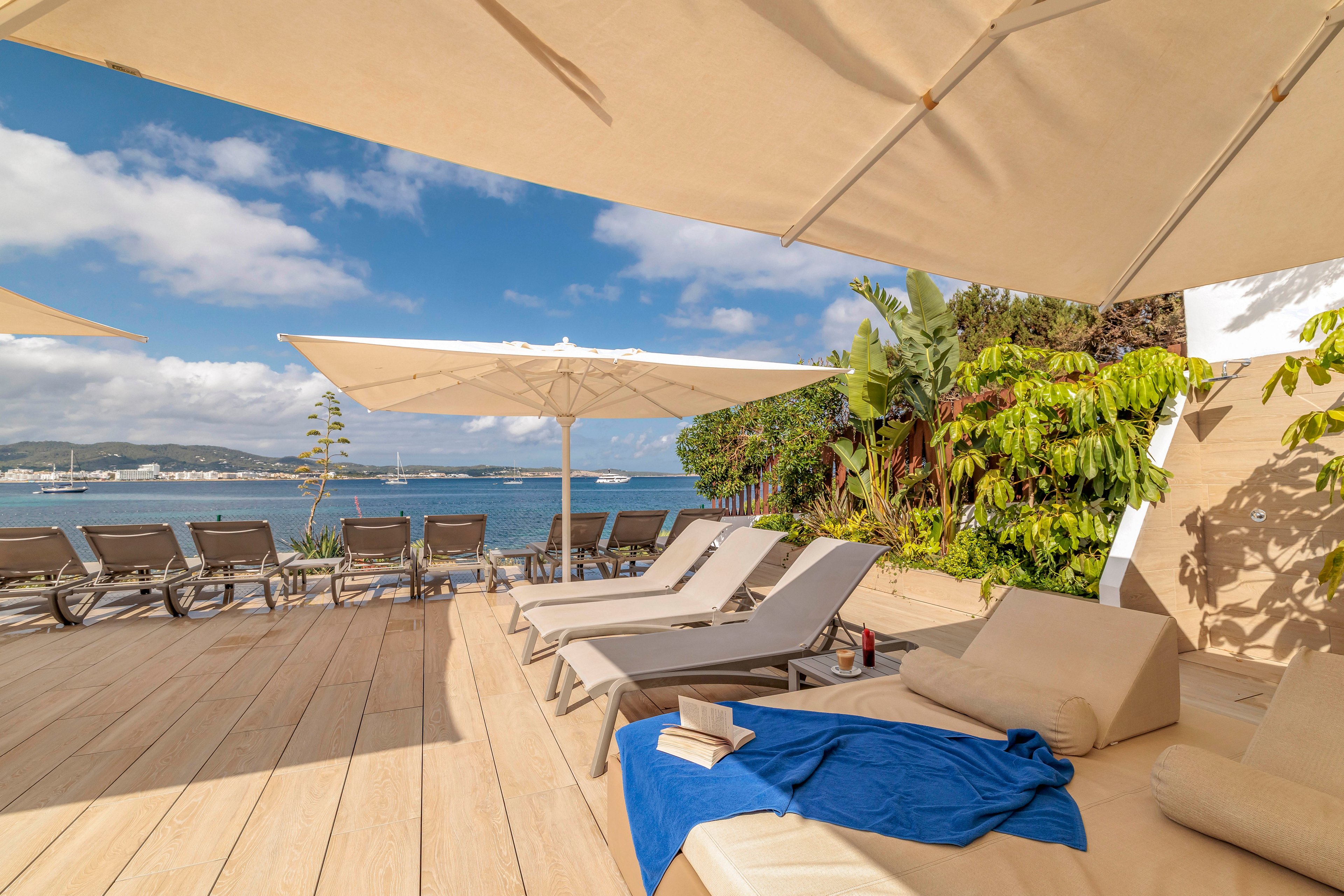 A bright seaside terrace features several cushioned lounge chairs arranged under large white sun umbrellas. The setting overlooks calm blue water with boats and a distant shoreline visible across the bay. A blue towel and an open book rest on one of the loungers, suggesting relaxation and leisure. Lush green plants line the edge of the deck, adding a tropical feel to the modern outdoor space.