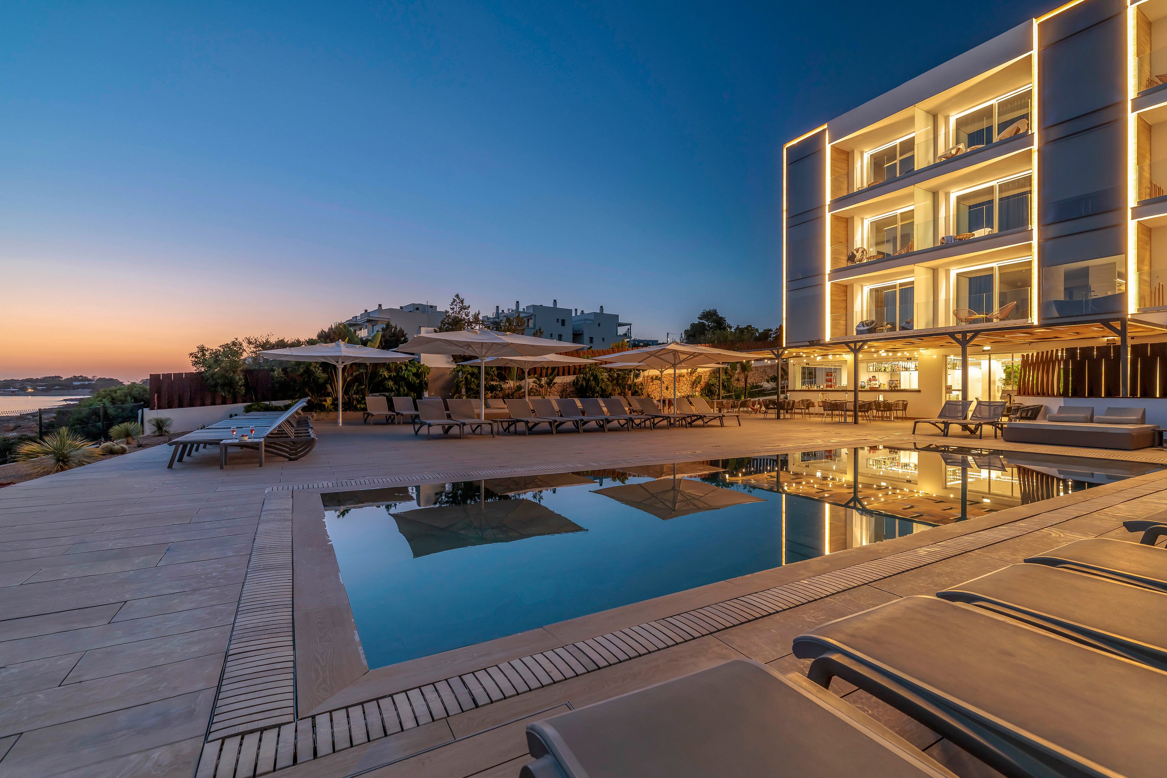 A contemporary multi-story hotel building is illuminated at dusk beside a calm outdoor swimming pool. Lounge chairs and sun umbrellas line the poolside terrace, facing the water and distant coastline. The sky transitions from warm orange near the horizon to deep blue, reflecting softly in the pool. The scene conveys a tranquil vacation atmosphere with clean architectural lines and subtle lighting.
