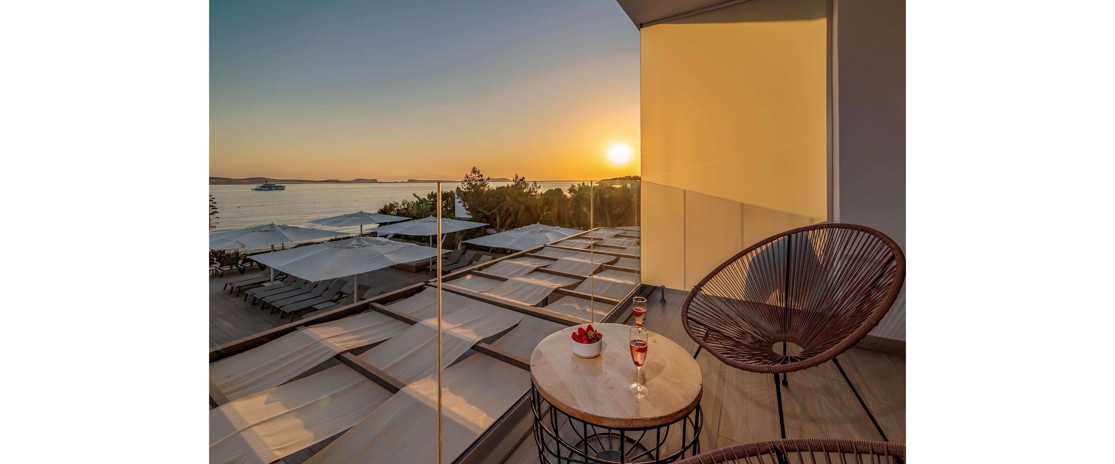A modern terrace overlooks a calm sea at sunset, with warm golden light reflecting off glass and metal surfaces. In the foreground, a small round table holds a glass of red drink and a bowl of strawberries beside a contemporary wire-frame chair. Below the balcony, rows of sun loungers and large white parasols line the waterfront deck. The scene conveys a relaxed vacation atmosphere with minimalist, stylish outdoor furniture and a serene coastal horizon.
