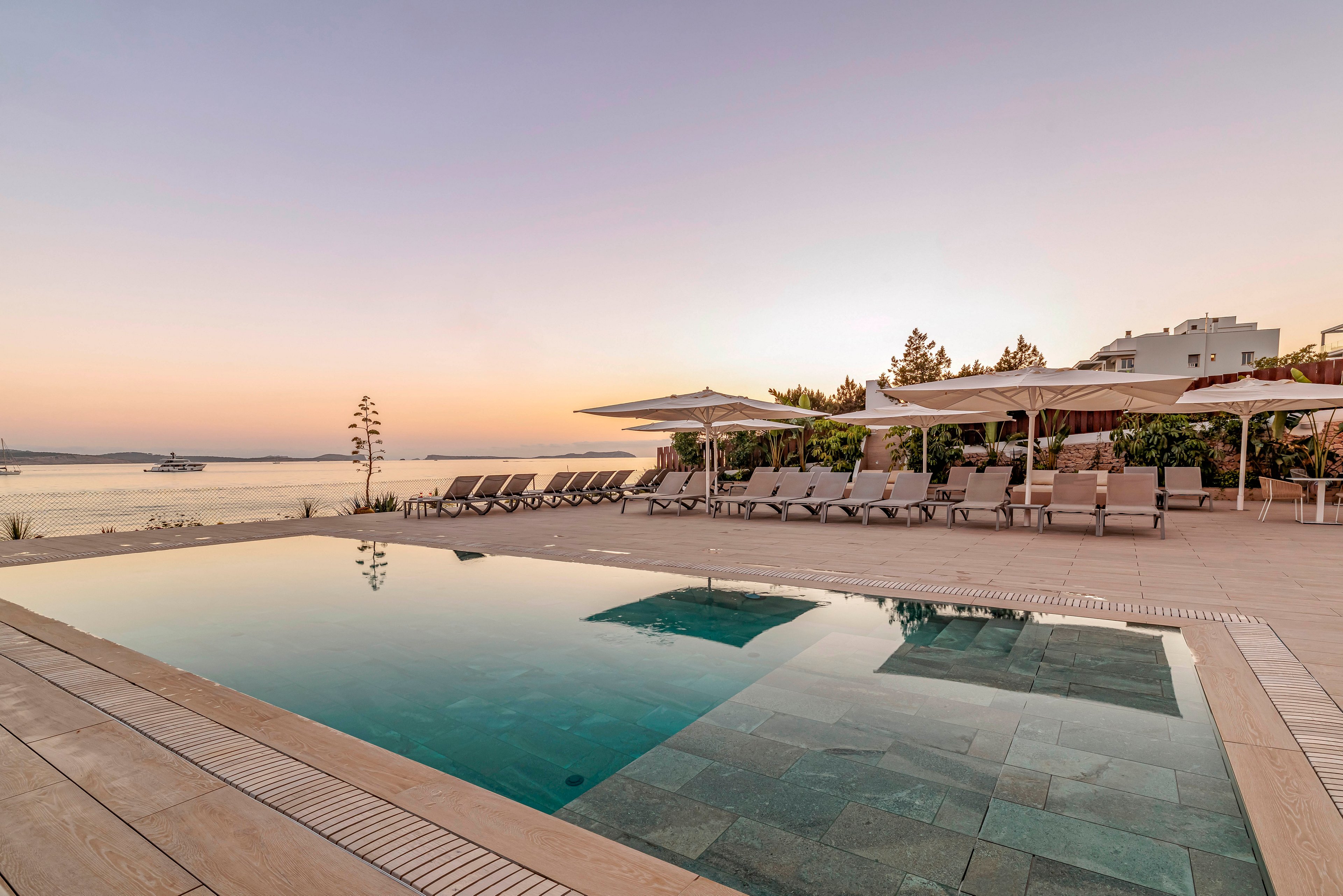 An outdoor swimming pool sits beside a row of sun loungers under large white umbrellas on a coastal terrace. The calm sea and distant shoreline are visible beyond the pool, bathed in soft evening light. The sky glows with pastel hues of pink and orange, creating a tranquil vacation atmosphere. Modern resort buildings and minimal landscaping frame the relaxing waterfront setting.