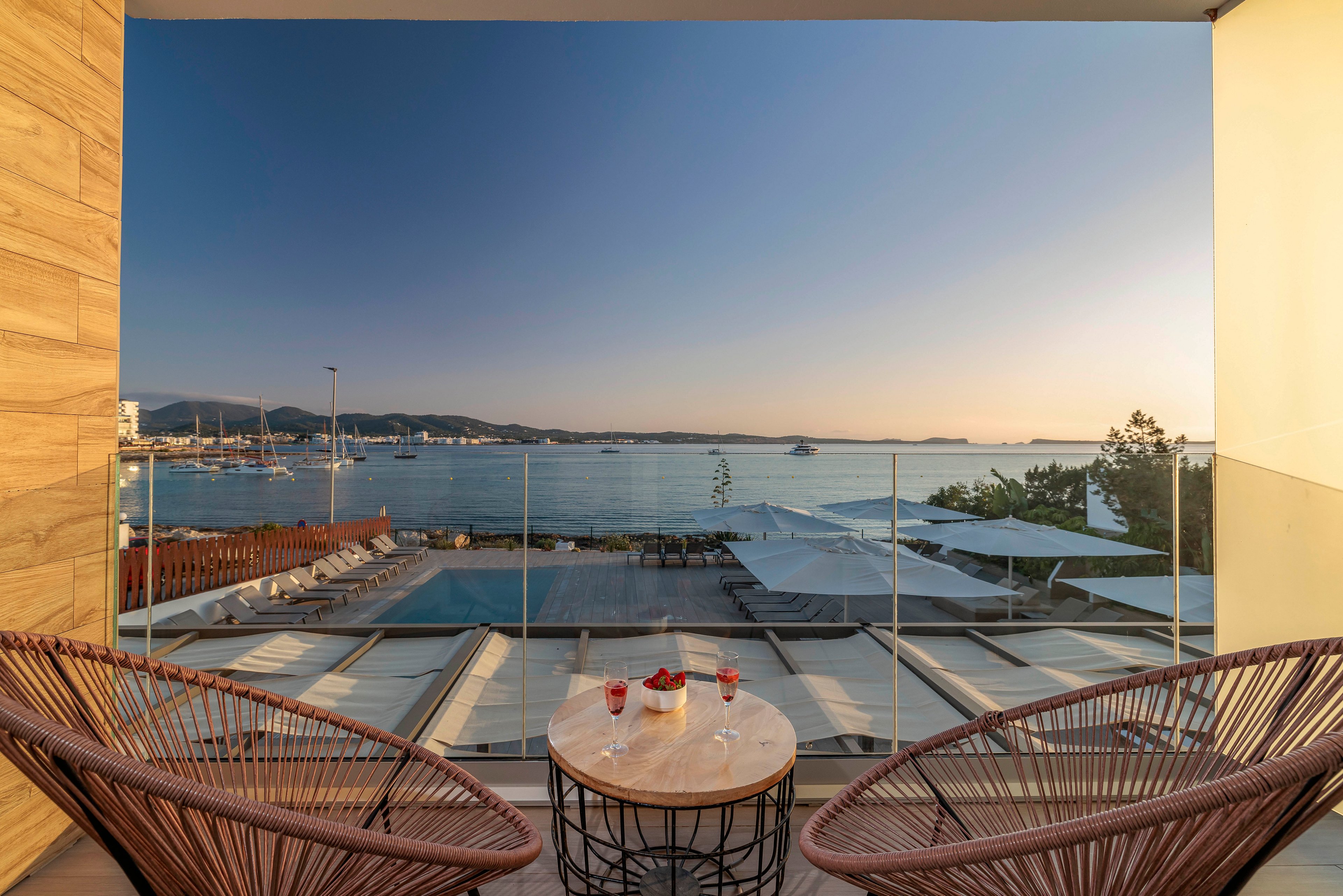 A modern terrace overlooks a calm coastal harbor at sunset. Two woven lounge chairs face a small round table with drinks, framed by glass railing and a pool area below. The sky transitions from warm tones near the horizon to clear blue above, reflecting softly on the water. Boats, distant hills, and waterfront structures complete the tranquil vacation setting.
