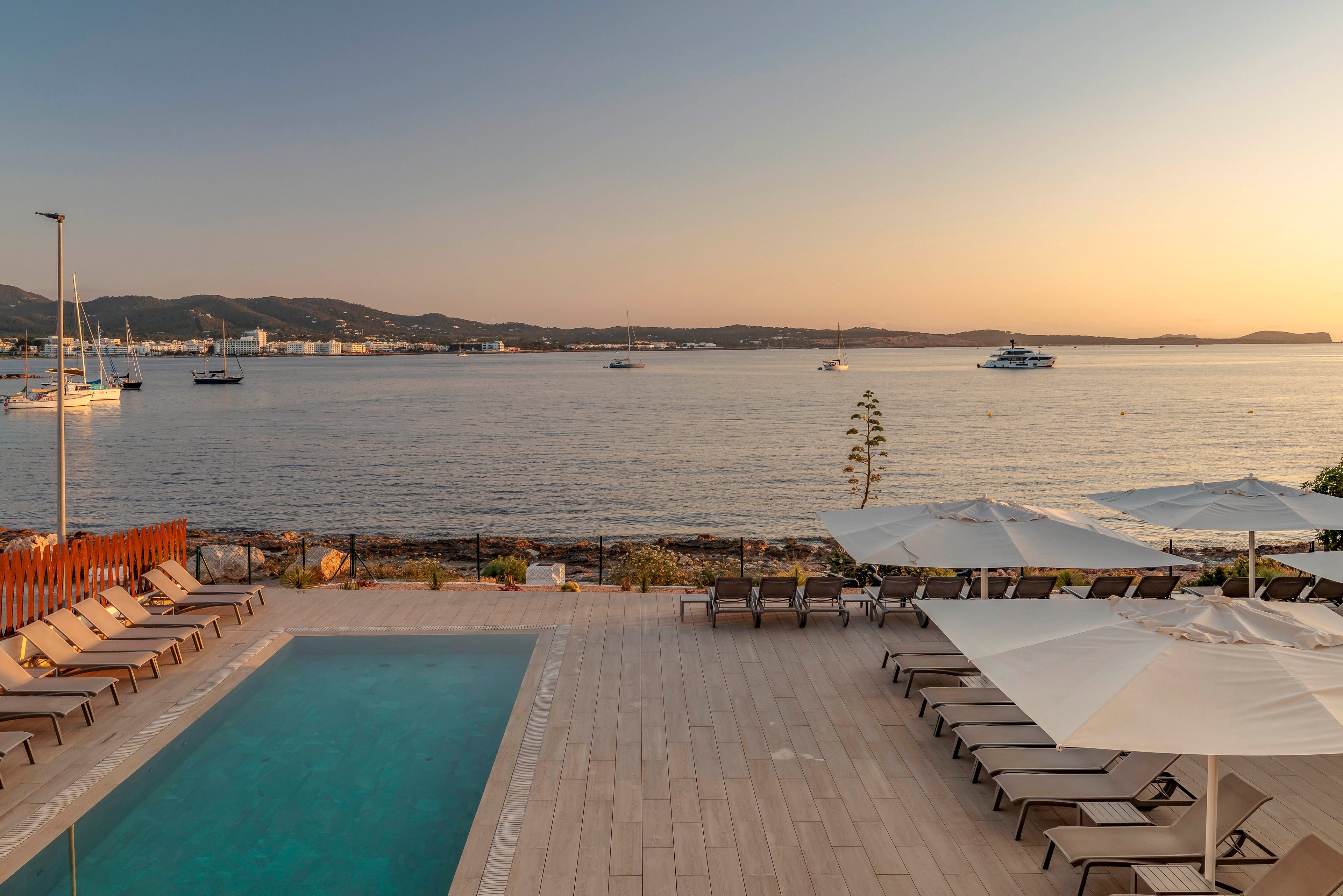 A rectangular outdoor swimming pool sits beside rows of empty lounge chairs and large white umbrellas at a seaside hotel. Beyond the pool area, calm water stretches out toward distant hills under a warm sunset sky. Several boats and yachts are anchored in the bay, adding to the relaxed coastal atmosphere. The soft golden light and clear weather create a peaceful vacation setting ideal for relaxation.