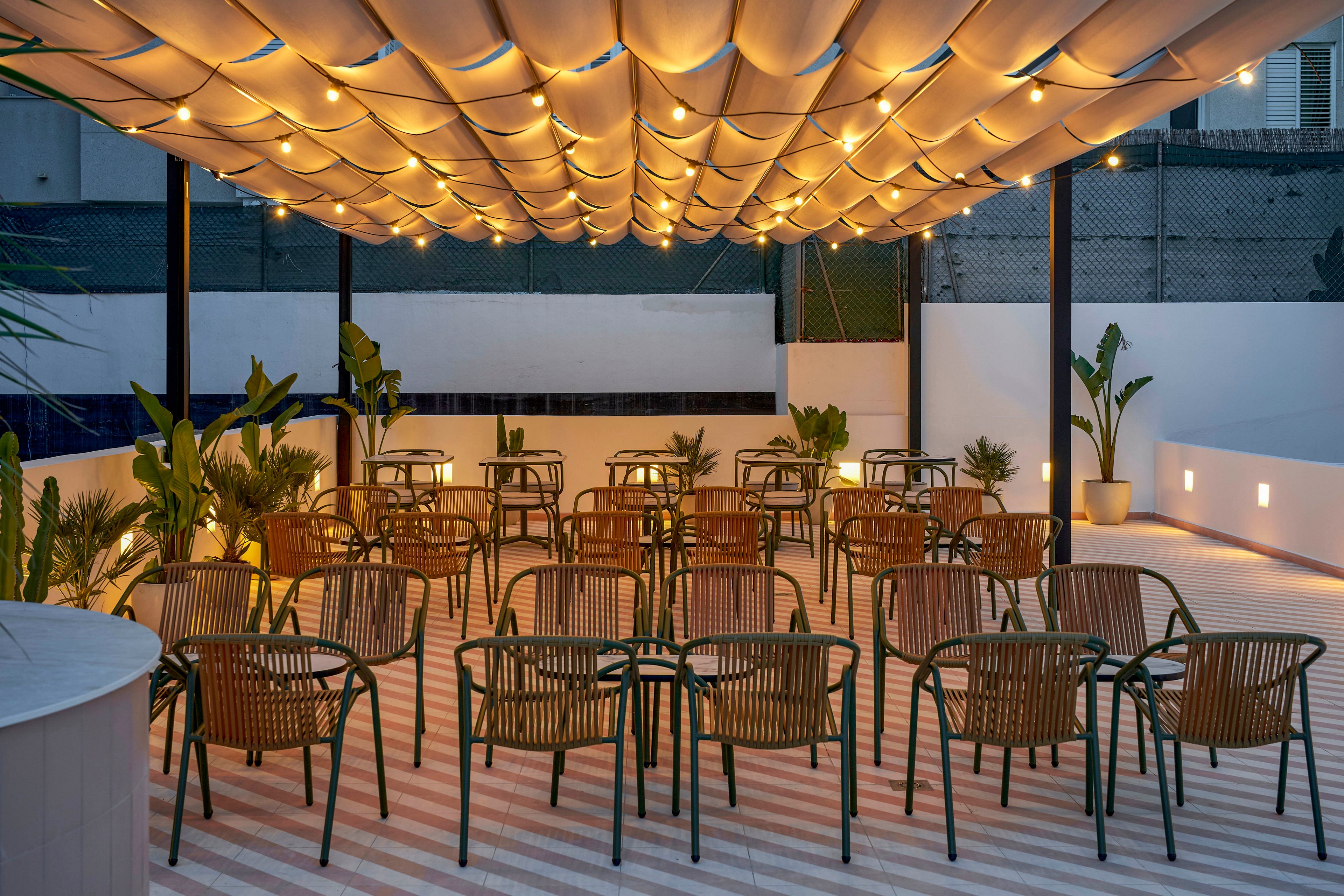 An open-air terrace is arranged with rows of empty chairs facing a small stage or focal area. Overhead, a fabric canopy with hanging string lights creates a warm, inviting glow. Potted plants line the sides of the space, adding greenery against the clean white walls and striped floor. The scene suggests a modern venue prepared for an evening event or gathering.