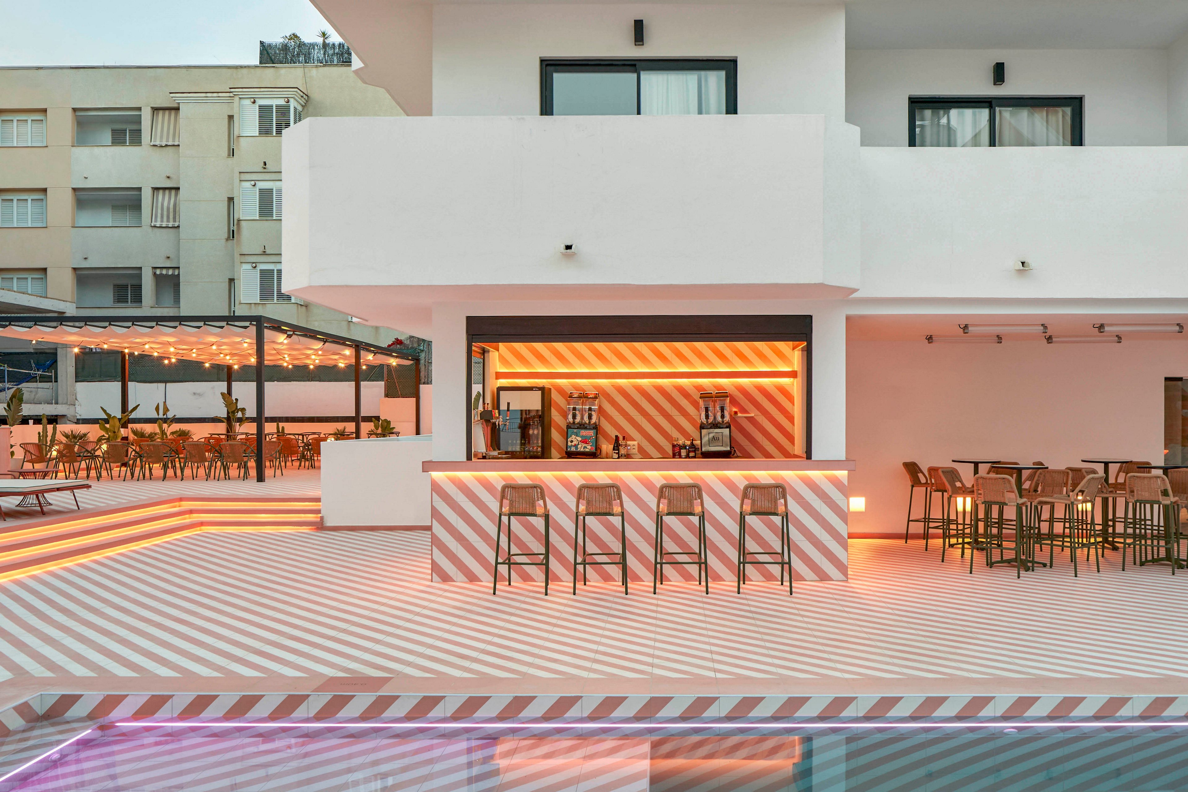 A stylish outdoor bar sits beside a swimming pool at a modern hotel. The architecture features clean white walls with geometric balconies and striped patterns on the bar front and pool deck. Bar stools and high tables create a relaxed social area, while warm accent lighting adds a welcoming glow. Nearby lounge chairs and covered seating suggest a resort atmosphere in an urban setting.