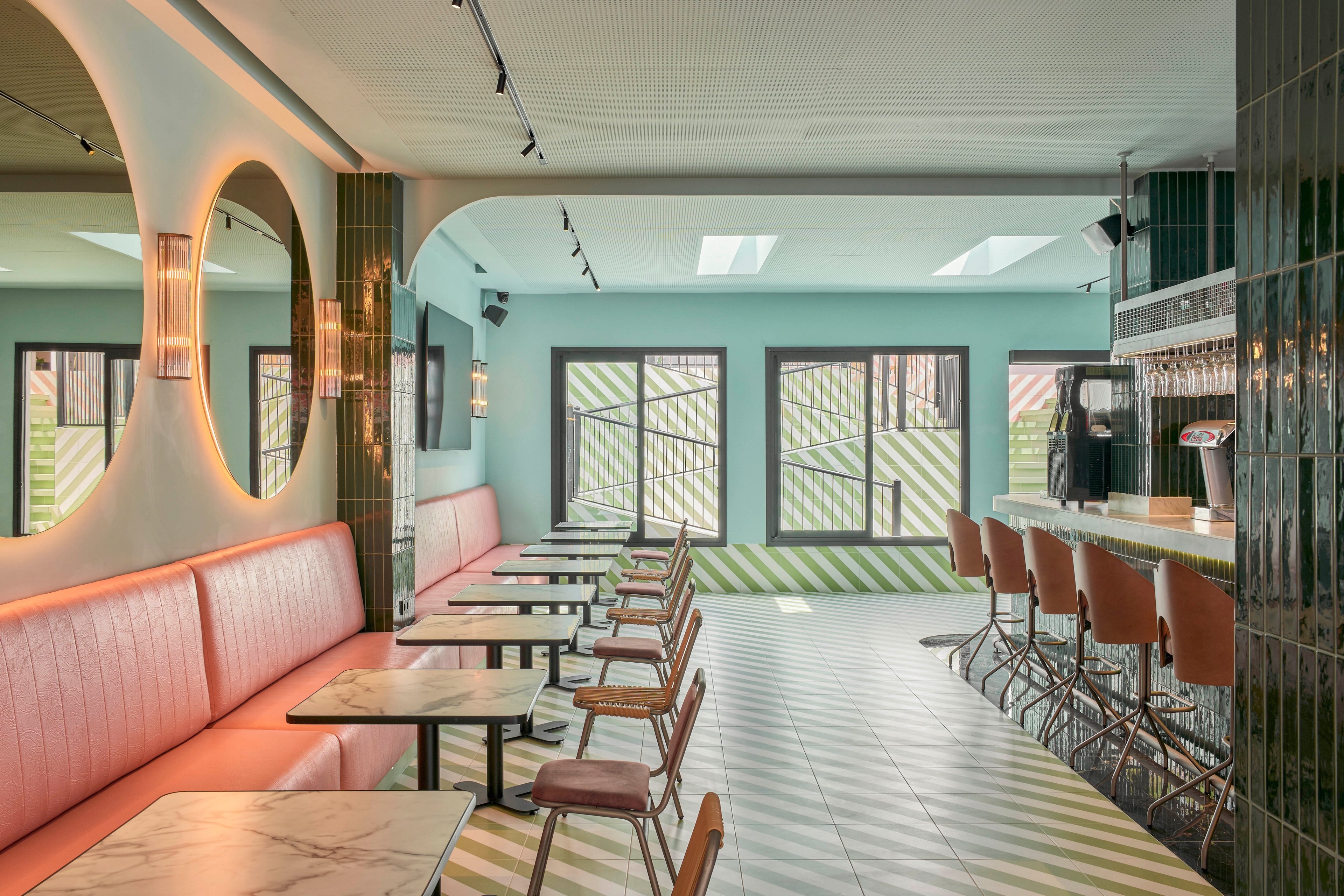 Contemporary cafe interior featuring pastel colors and clean lines. Pink upholstered banquette seating runs along one wall with small marble-top tables and wooden chairs. A long bar with matching pink barstools faces a row of windows and doors with geometric striped patterns outside. Soft indirect lighting, round mirrors, and tiled surfaces create a stylish, retro-inspired atmosphere in an empty space.