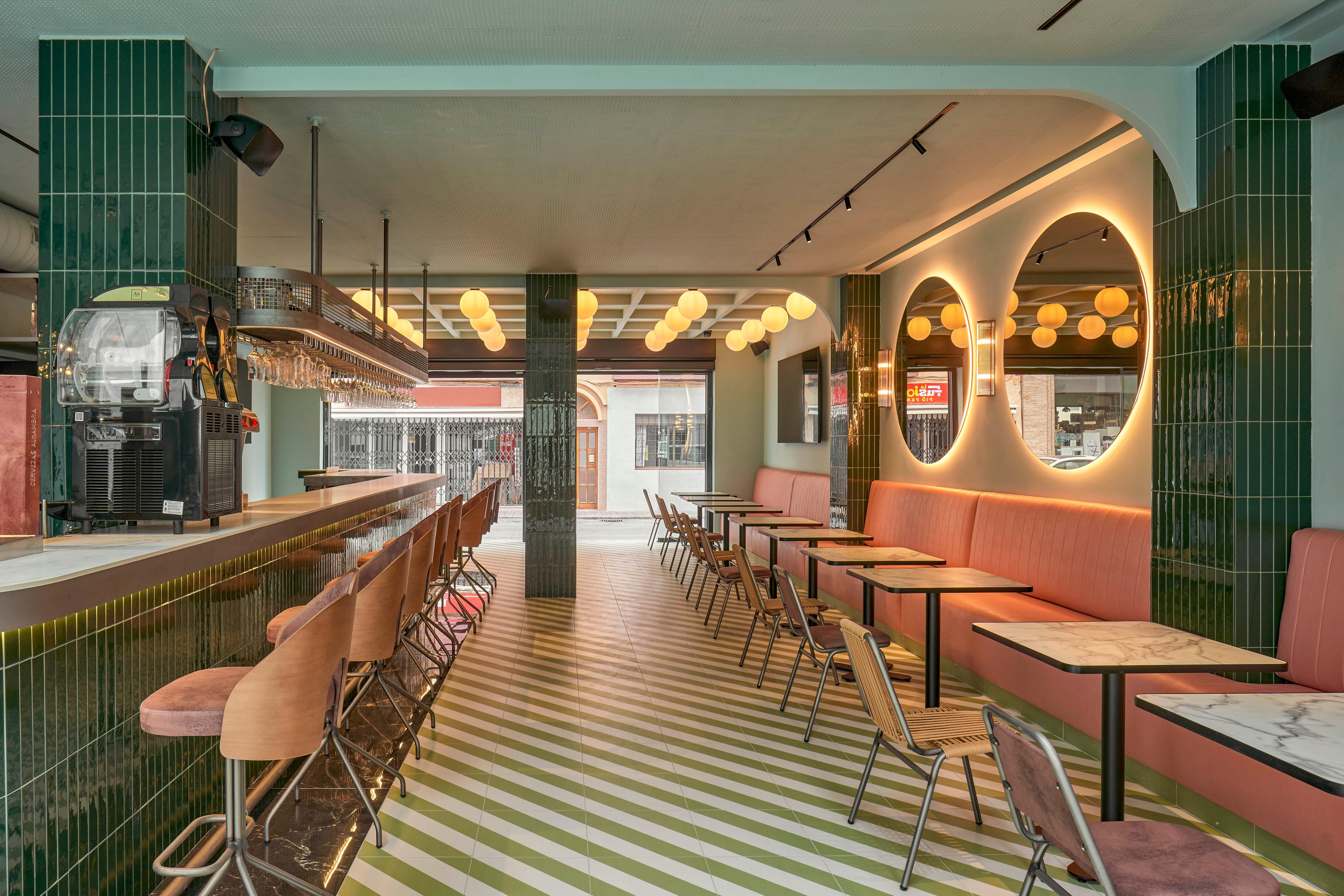 Contemporary bar interior featuring a long counter with upholstered bar stools and a row of small tables with chairs. The setting is bright and stylish, with green tiled walls, pink banquette seating, and a striped green-and-white floor. Circular mirrors and warm pendant lights create a cozy, inviting ambiance. Large front windows open to the street, adding natural light and an urban feel to the space.