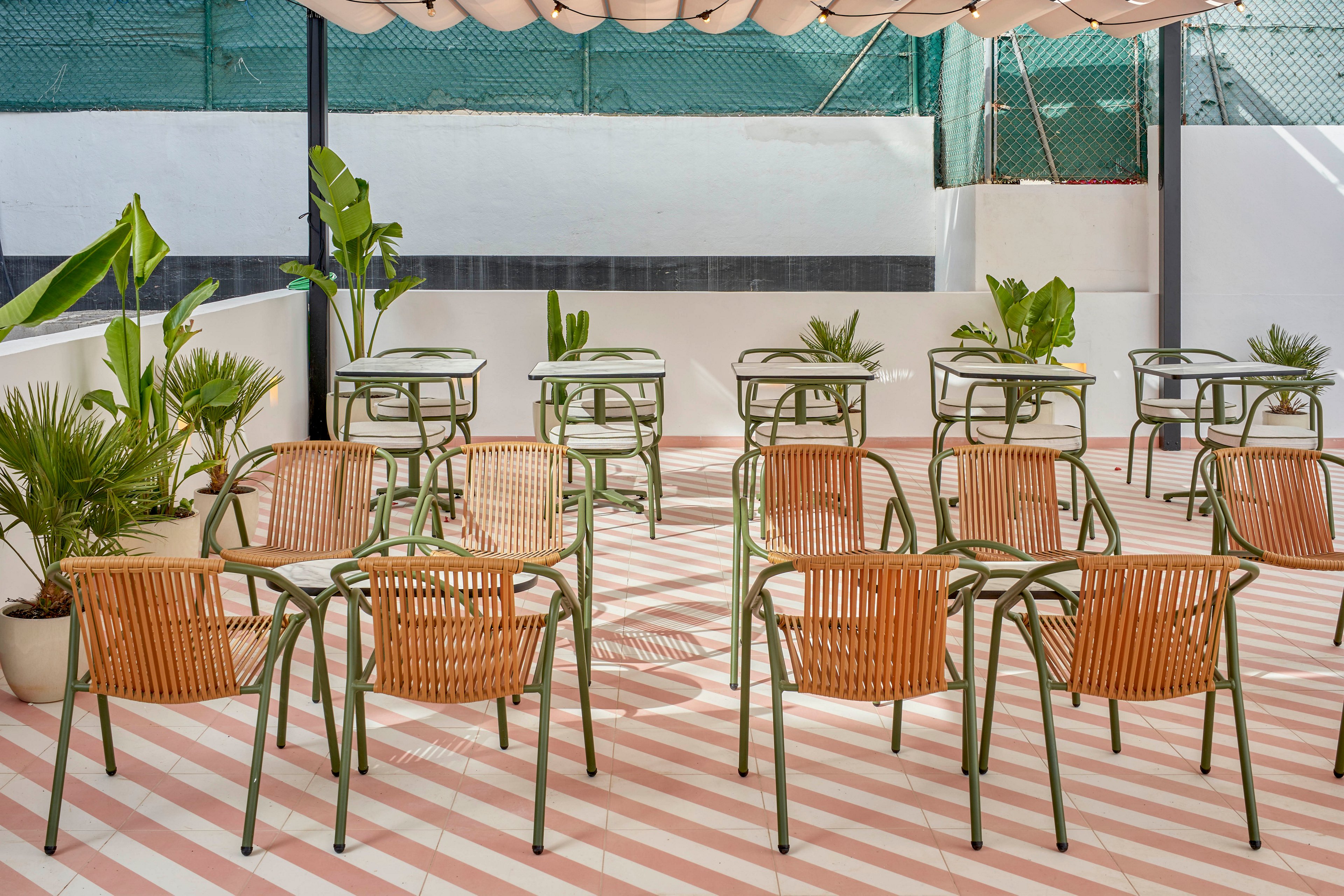 An empty outdoor seating area features rows of wicker chairs paired with green metal tables. The terrace floor has diagonal pink and white stripes that create a bold graphic pattern. Potted tropical plants are placed along the edges, adding greenery to the minimalist space. A canopy above and high walls around the terrace suggest a private, relaxed setting.