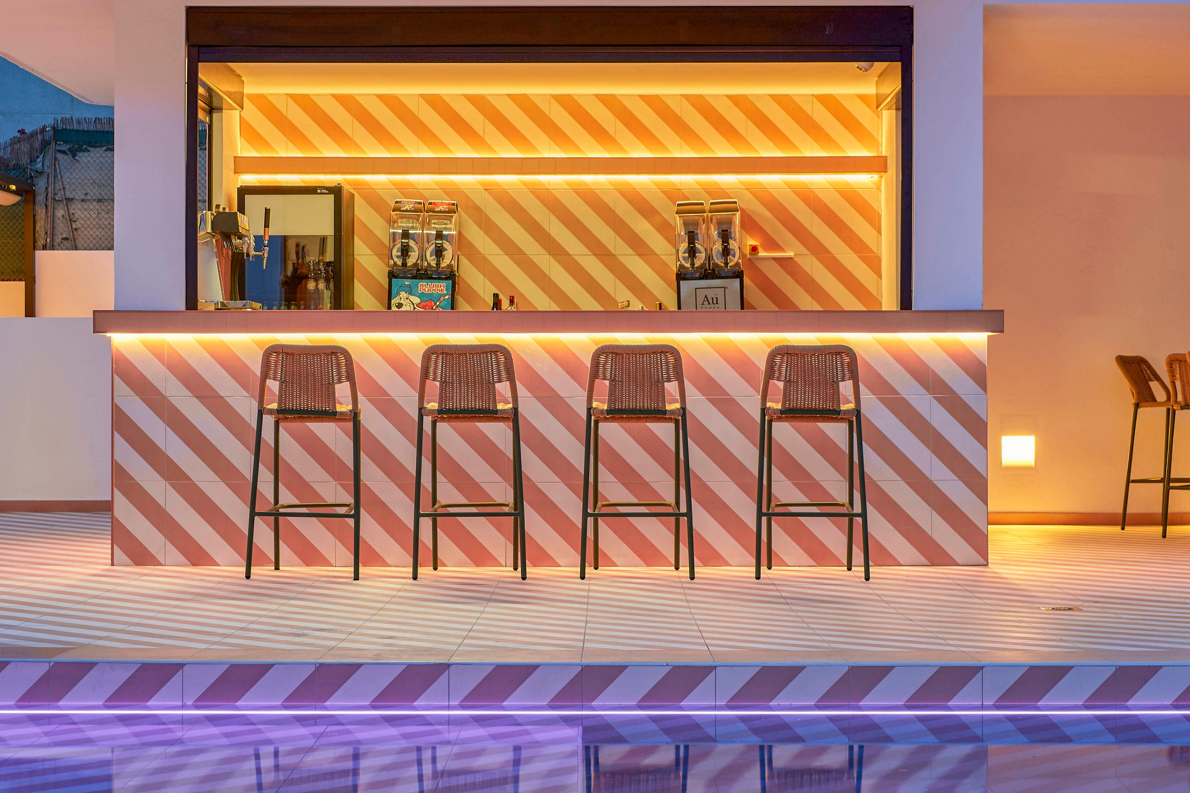 A contemporary outdoor bar with a striped illuminated counter stands beside a calm swimming pool. Four bar stools are neatly lined up in front of the counter, with warm ambient lighting creating a welcoming atmosphere. The geometric stripes on the bar and floor are reflected in the still pool water, enhancing the modern design. The setting appears tranquil and stylish, ideal for evening relaxation at a resort or hotel.