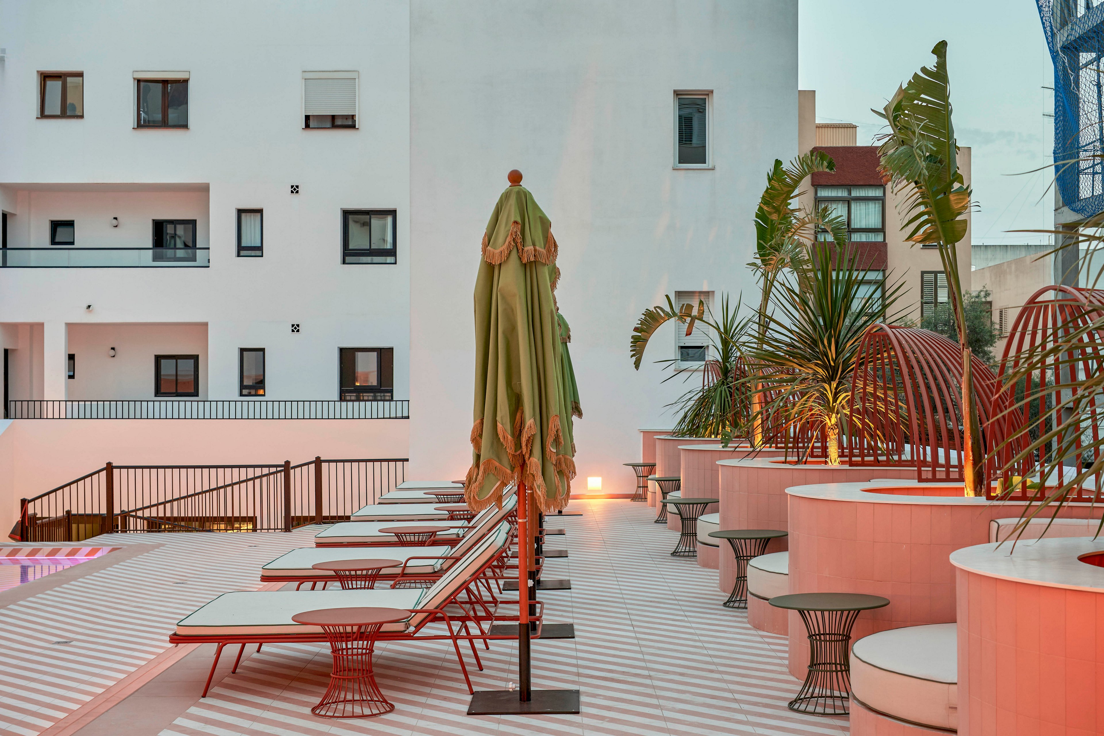 A contemporary rooftop terrace features a row of sun loungers and closed green umbrellas beside a pool. The setting is urban, surrounded by modern apartment buildings with simple facades and windows. Warm ambient lighting and soft evening sky tones create a relaxed atmosphere. Potted palm trees and curved seating elements add a stylish, resort-like feel to the outdoor space.