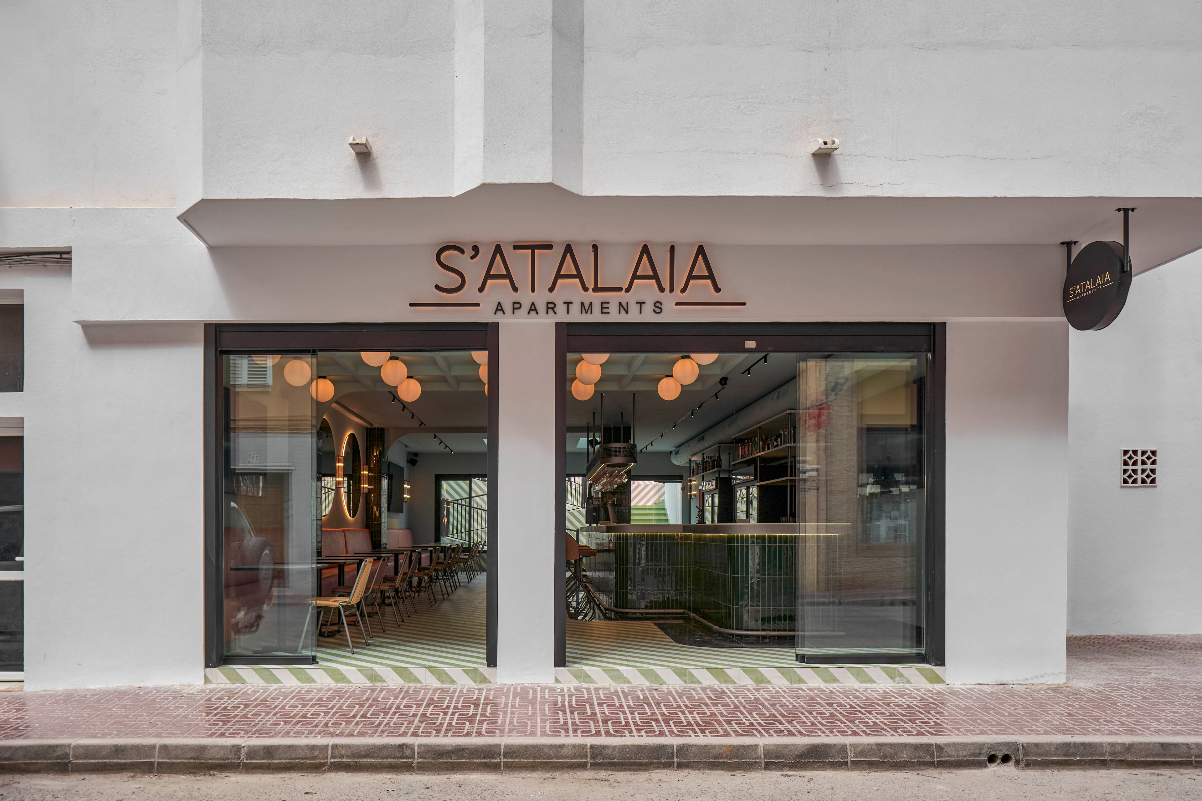 Street-level view of the S'Atalaia Apartments entrance shows a contemporary white facade with large glass windows. Warm interior lighting and hanging globe lamps reveal a stylish lobby and seating area inside. The exterior features clean lines, a subtle striped floor pattern at the threshold, and a narrow sidewalk in front. A small round sign projects from the wall, reinforcing the apartment name for passersby.