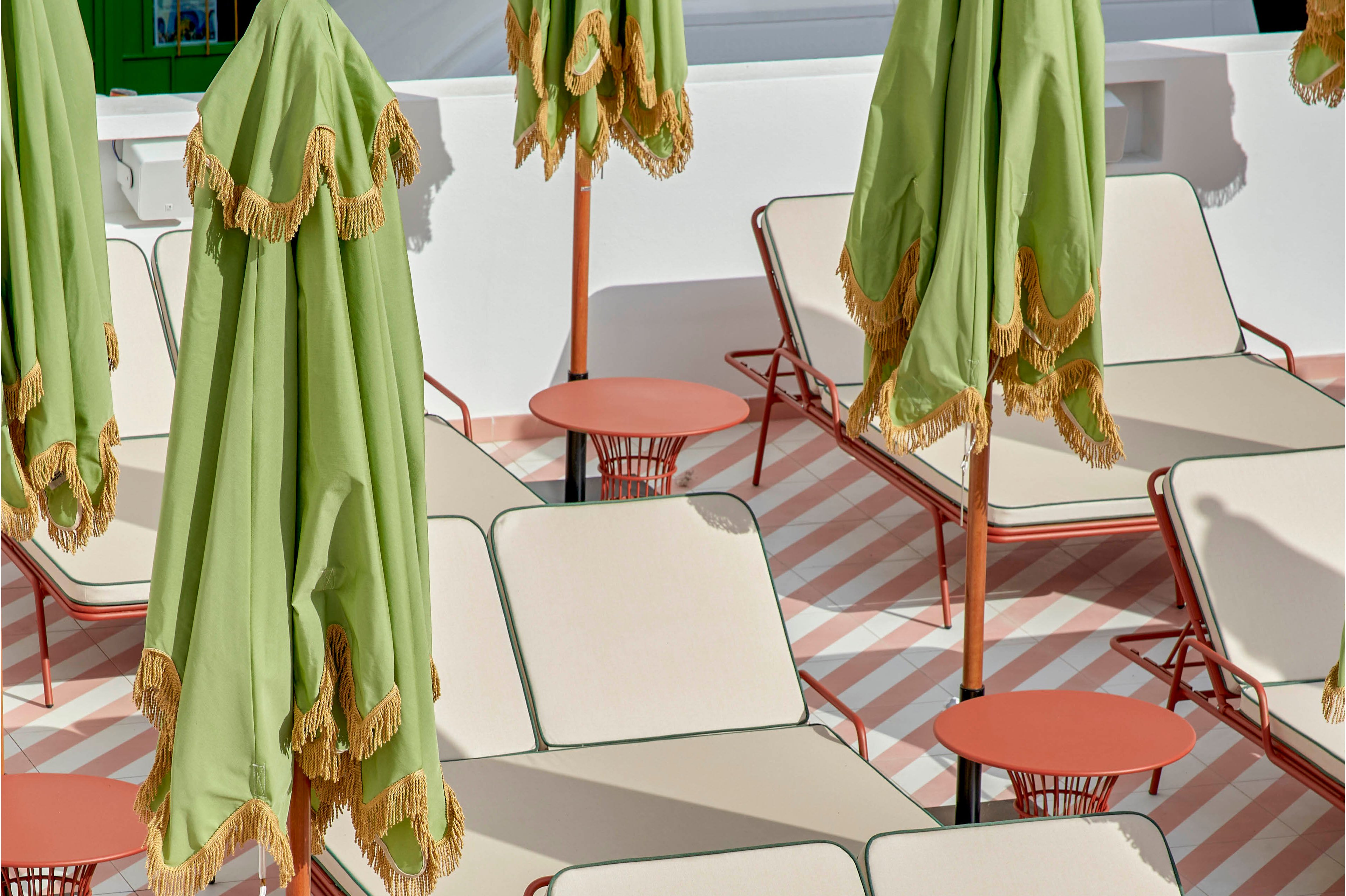 A group of cream-colored sun loungers is arranged neatly on a striped patio. Tall green umbrellas with decorative fringe stand between the loungers, casting shade over the seating area. Small round red side tables accompany each lounger, adding contrast to the soft pink and white striped floor. The scene suggests a stylish, modern outdoor pool or terrace area with no people present.
