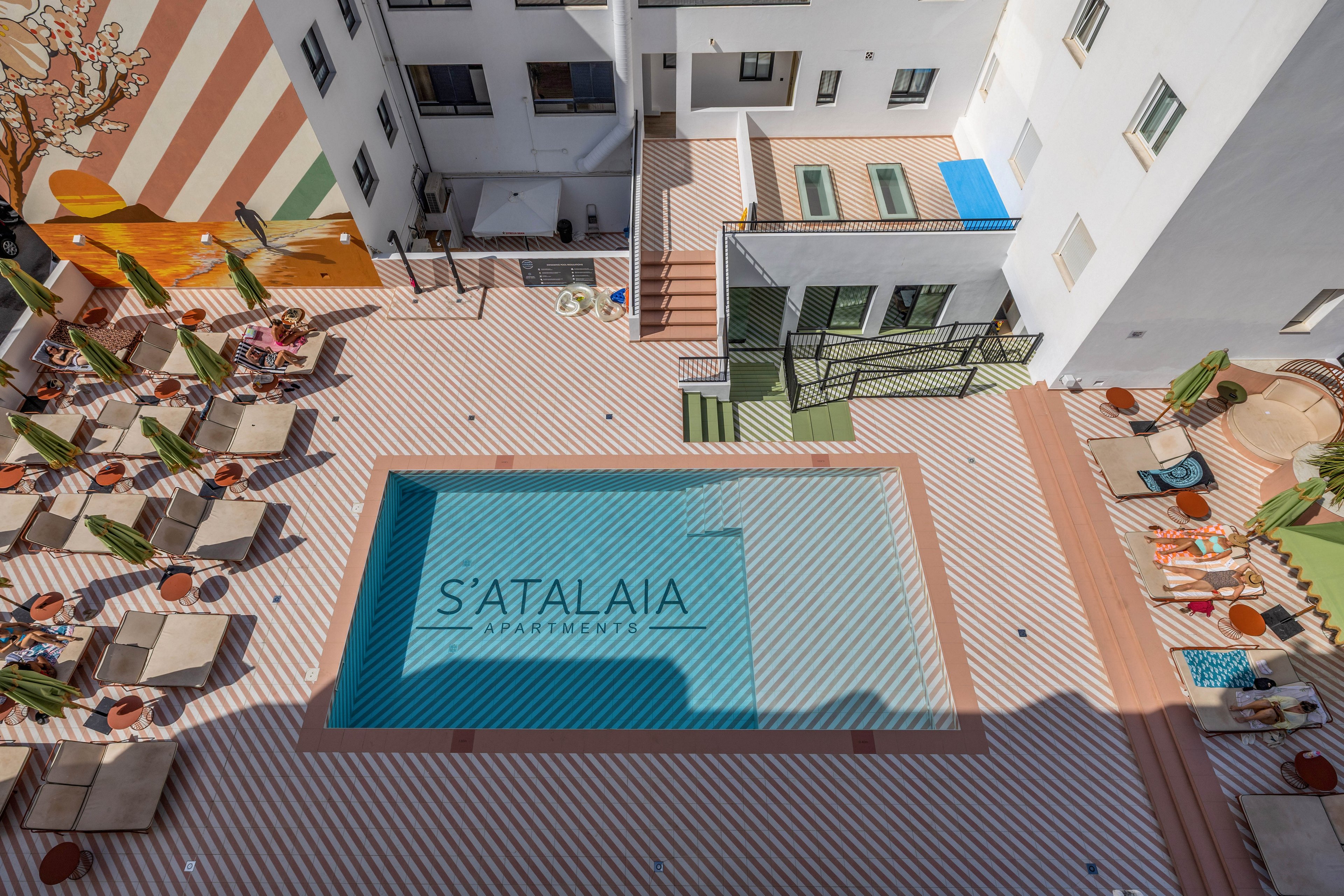 A rectangular outdoor swimming pool sits at the center of a modern hotel courtyard viewed from above. The pool has the text "S'ATALAIA" visible on its floor. Around the pool are neatly arranged sun loungers with umbrellas and small tables, some with towels and drinks. The striped tile pattern and white building walls create a clean, contemporary aesthetic. Bright sunlight casts strong shadows, emphasizing the geometric layout of the space.