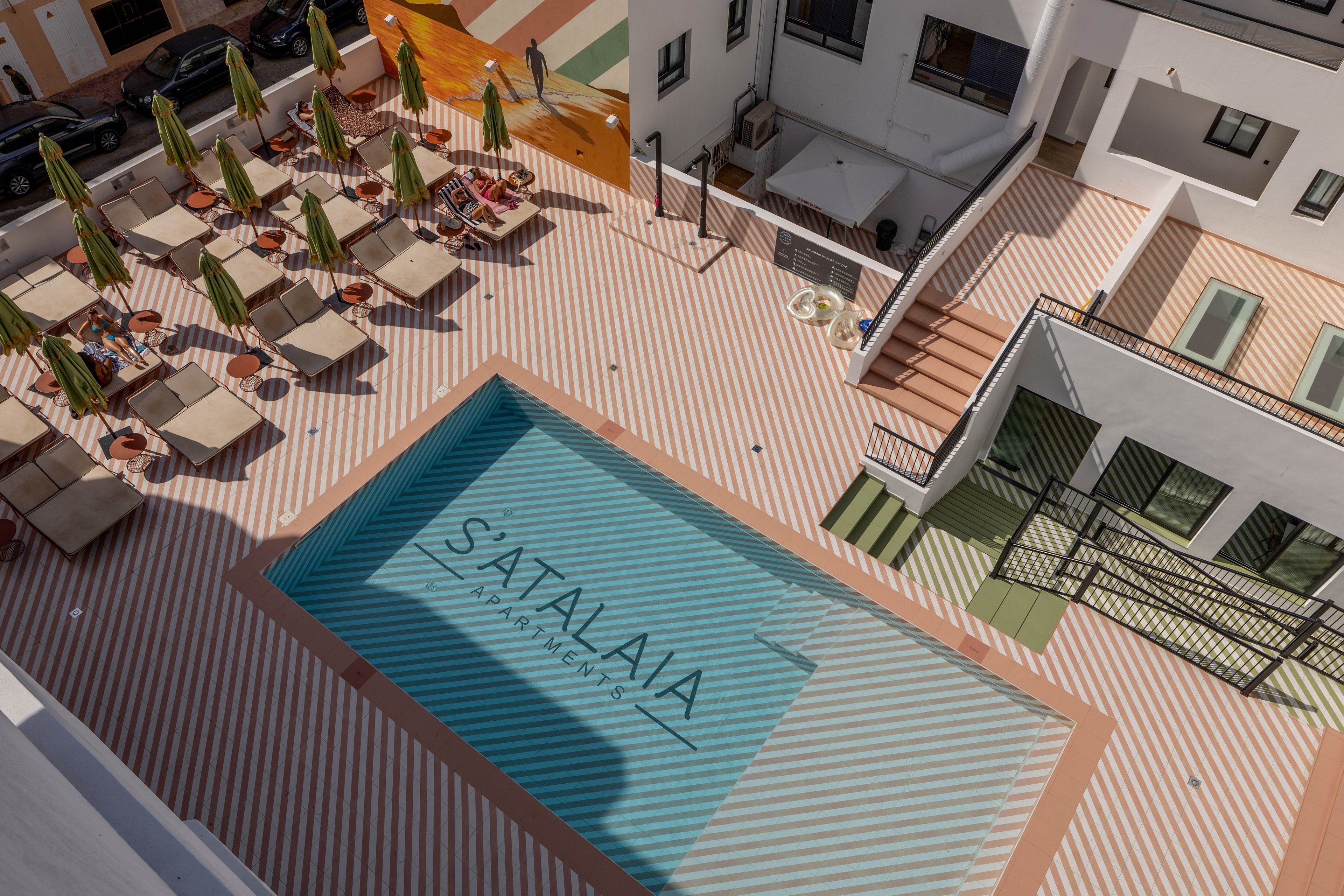A contemporary hotel courtyard is viewed from above, featuring a rectangular swimming pool at its center. The pool floor displays the text "SATA ALAIA" along with smaller wording beneath it. Surrounding the water are neatly arranged sun loungers with umbrellas and small side tables. The striped deck pattern and clean white building facades create a stylish, geometric look. Stairways, railings, and upper-level walkways frame the open-air relaxation area.