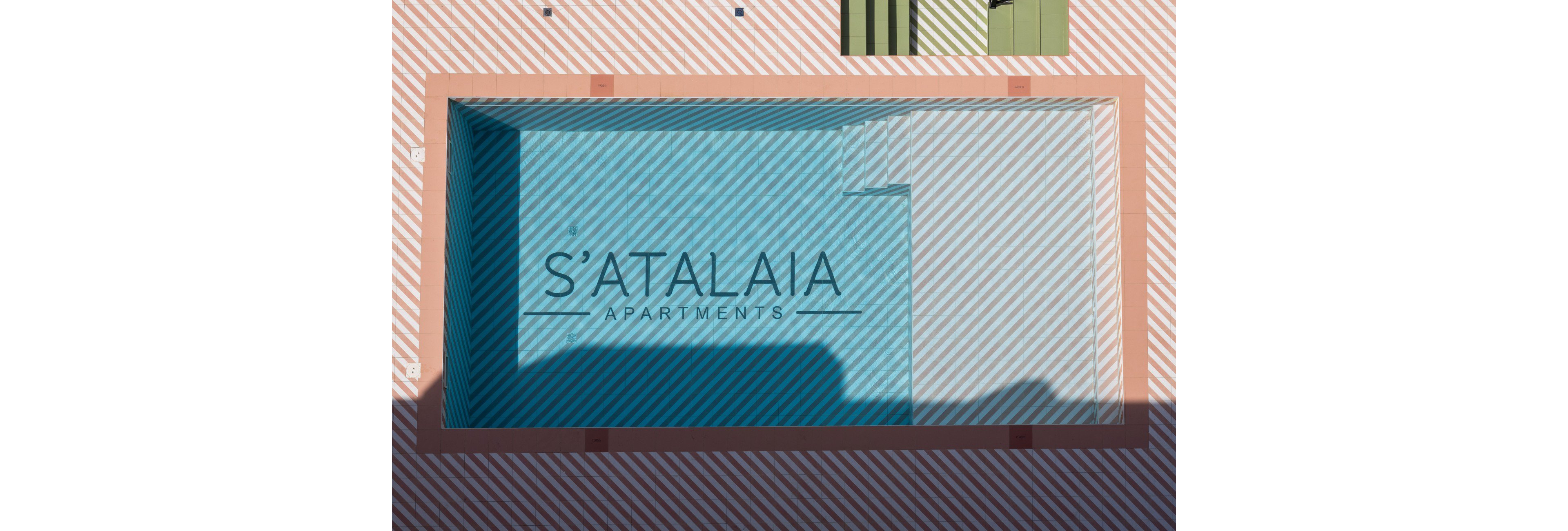 Top-down view of an empty rectangular swimming pool at S'Atalaia Apartments. The pool interior is blue with diagonal white stripes, framed by a muted pink border. Shadows fall across part of the pool, adding contrast and depth. The words "S'ATALAIA APARTMENTS" are clearly visible in the center of the pool.