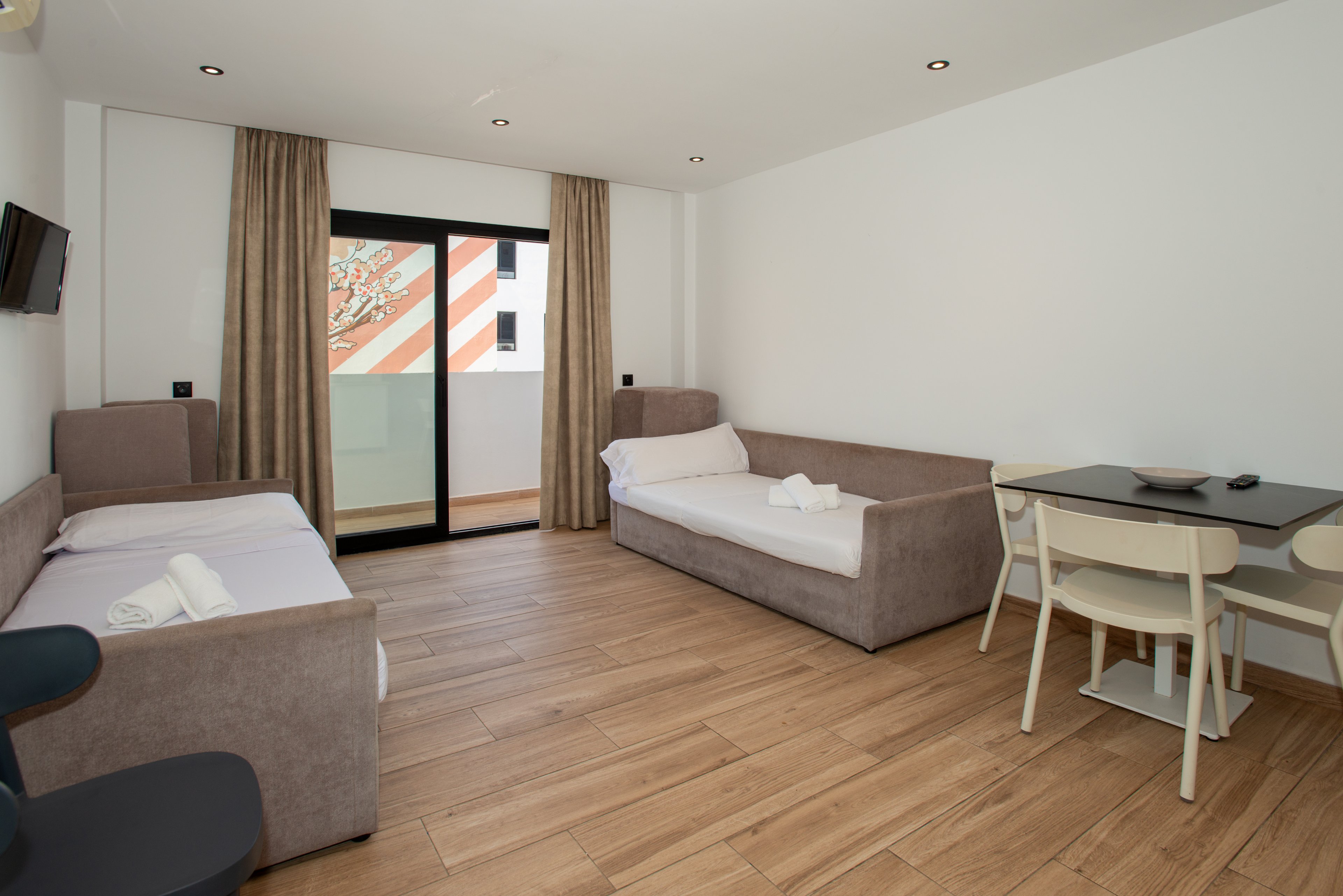 Bright modern hotel room featuring two single beds with folded white towels. The space has light wood flooring, neutral-colored sofas, and simple furnishings. A small dining table with chairs sits near the wall opposite a wall-mounted television. Large sliding glass doors lead to a balcony with a view of a neighboring building with striped exterior decor. Recessed ceiling lights and natural daylight create a clean, minimalist atmosphere.