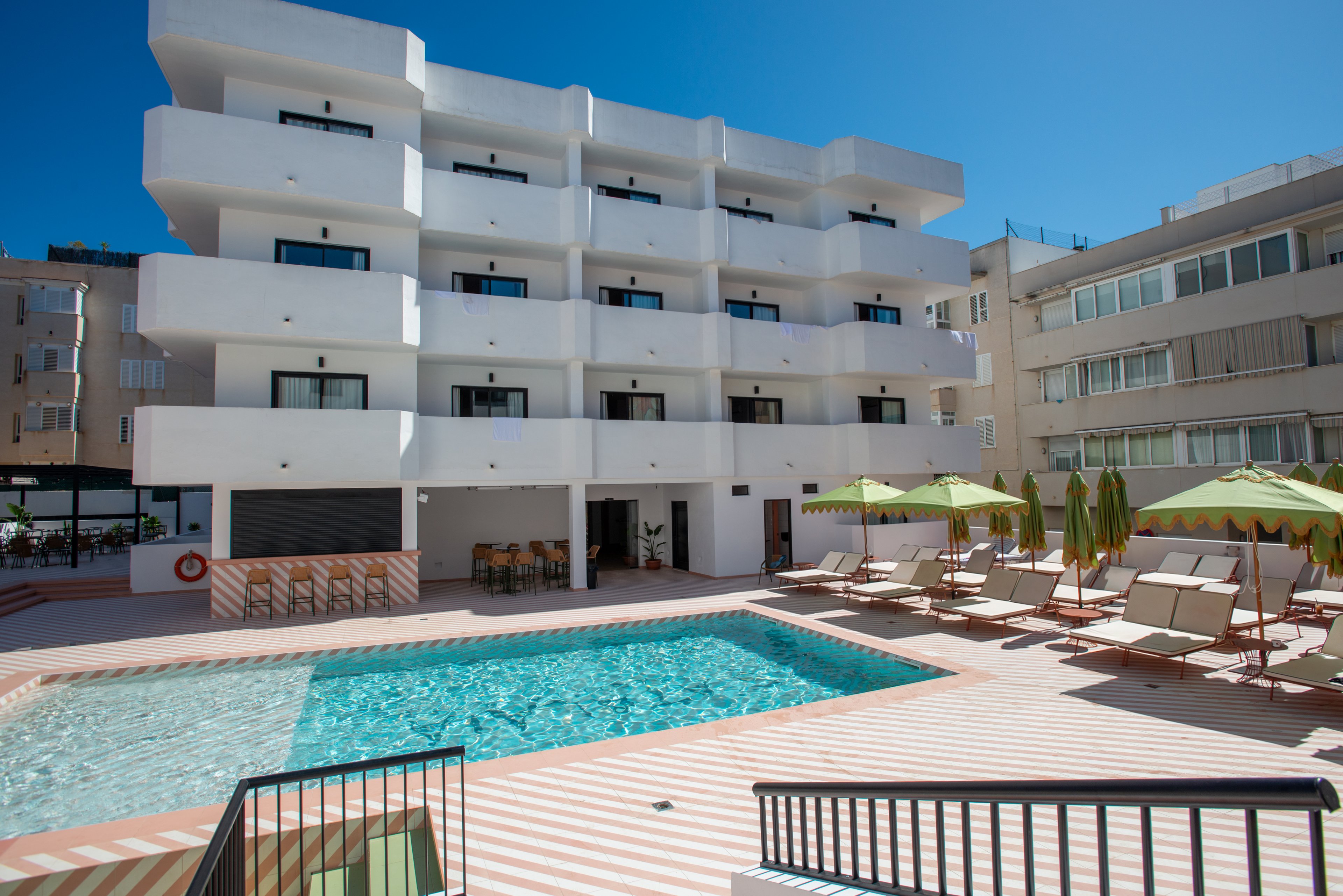 A modern white hotel building overlooks a clear blue outdoor swimming pool. Sun loungers and green umbrellas line the poolside terrace, suggesting a relaxing vacation setting. The sky is bright and cloudless, emphasizing the warm, sunny weather. Surrounding urban buildings frame the courtyard, giving the scene a contemporary resort-in-the-city feel.