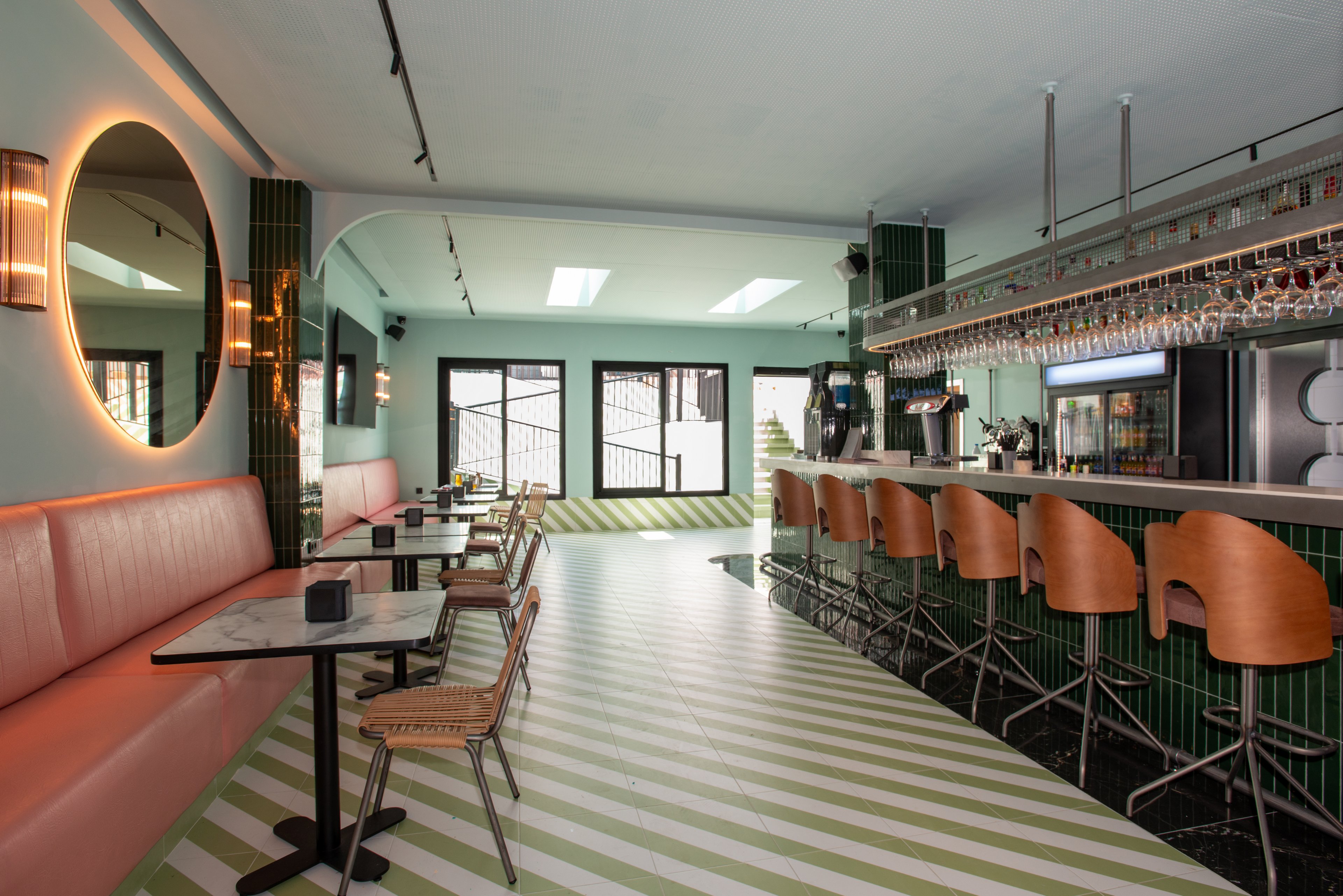 Contemporary bar interior featuring a long counter with wooden bar stools and a row of hanging glasses. On the left side, a cushioned pink banquette with small marble tables and wicker chairs lines the wall. The space is accented by a large round mirror, vertical green tiles, and a mint-green ceiling and walls. Diagonal green and white stripes cover the floor, leading toward large windows and doors at the back. The overall atmosphere is bright, stylish, and inviting, suitable for socializing or casual dining.