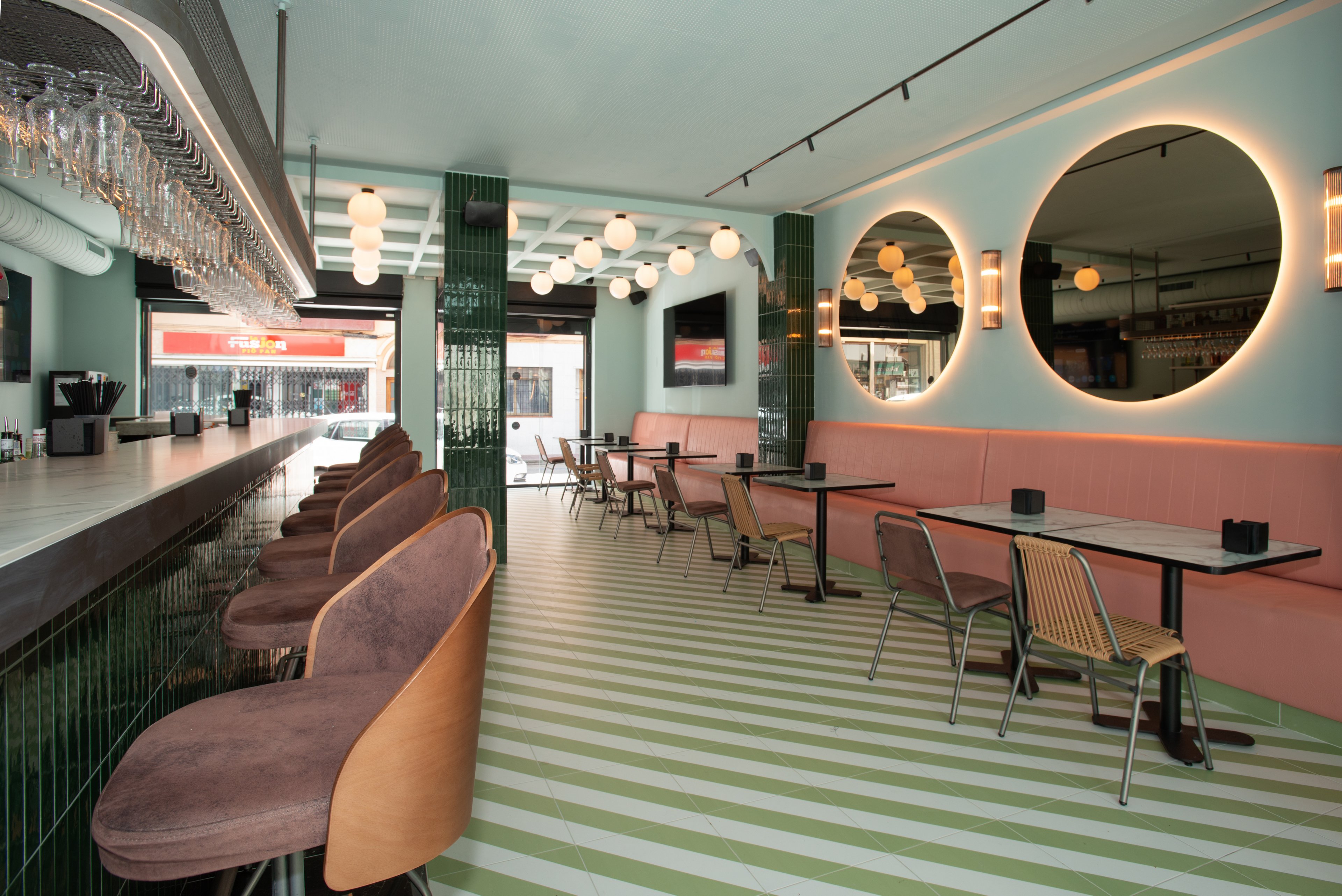 Contemporary bar interior featuring a long counter with upholstered stools on the left and small dining tables on the right. The space is decorated in pastel pink and green tones with glossy green columns and a striped floor. Large round mirrors with backlighting and globe pendant lights create a warm, inviting atmosphere. The bar appears empty, emphasizing the clean lines and modern design details.