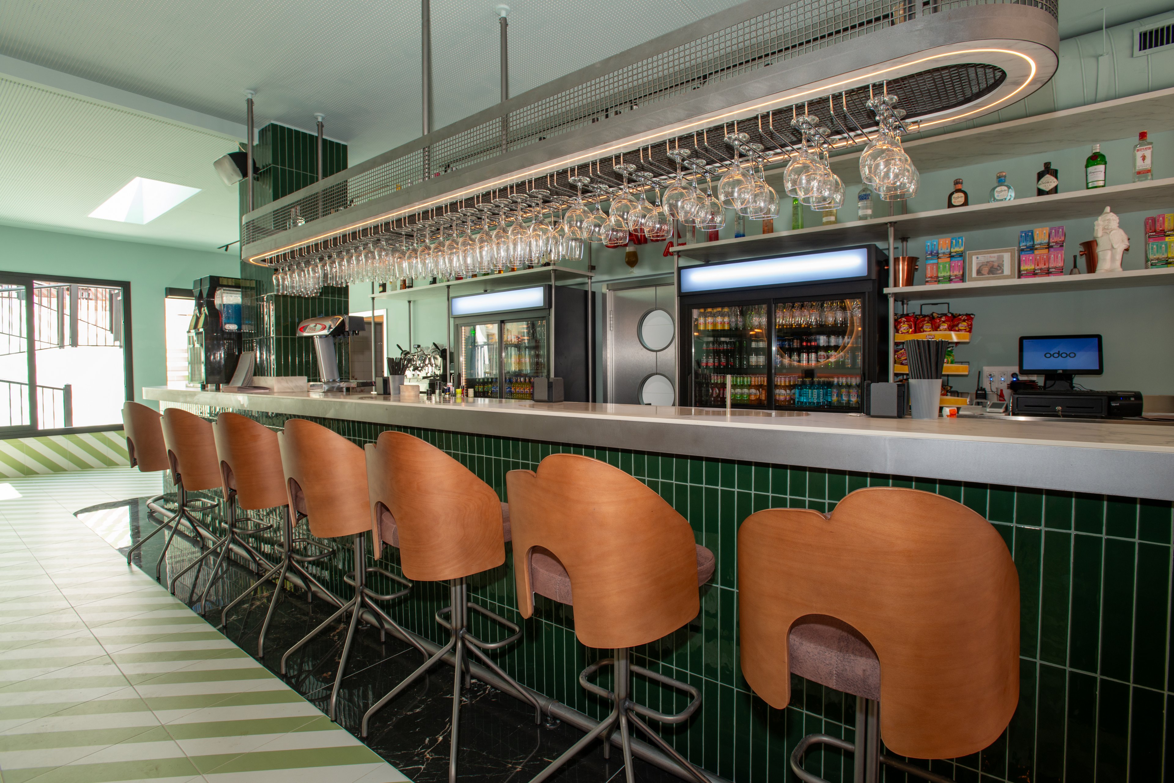 A contemporary bar interior features a long counter lined with wooden bar stools and green tiled fronts. Overhead racks display rows of hanging glassware above the bar. Refrigerated display cases and shelves stocked with bottles and snacks are visible behind the counter. The space is brightly lit with natural light from large windows and has a clean, minimalist design with green and white flooring.