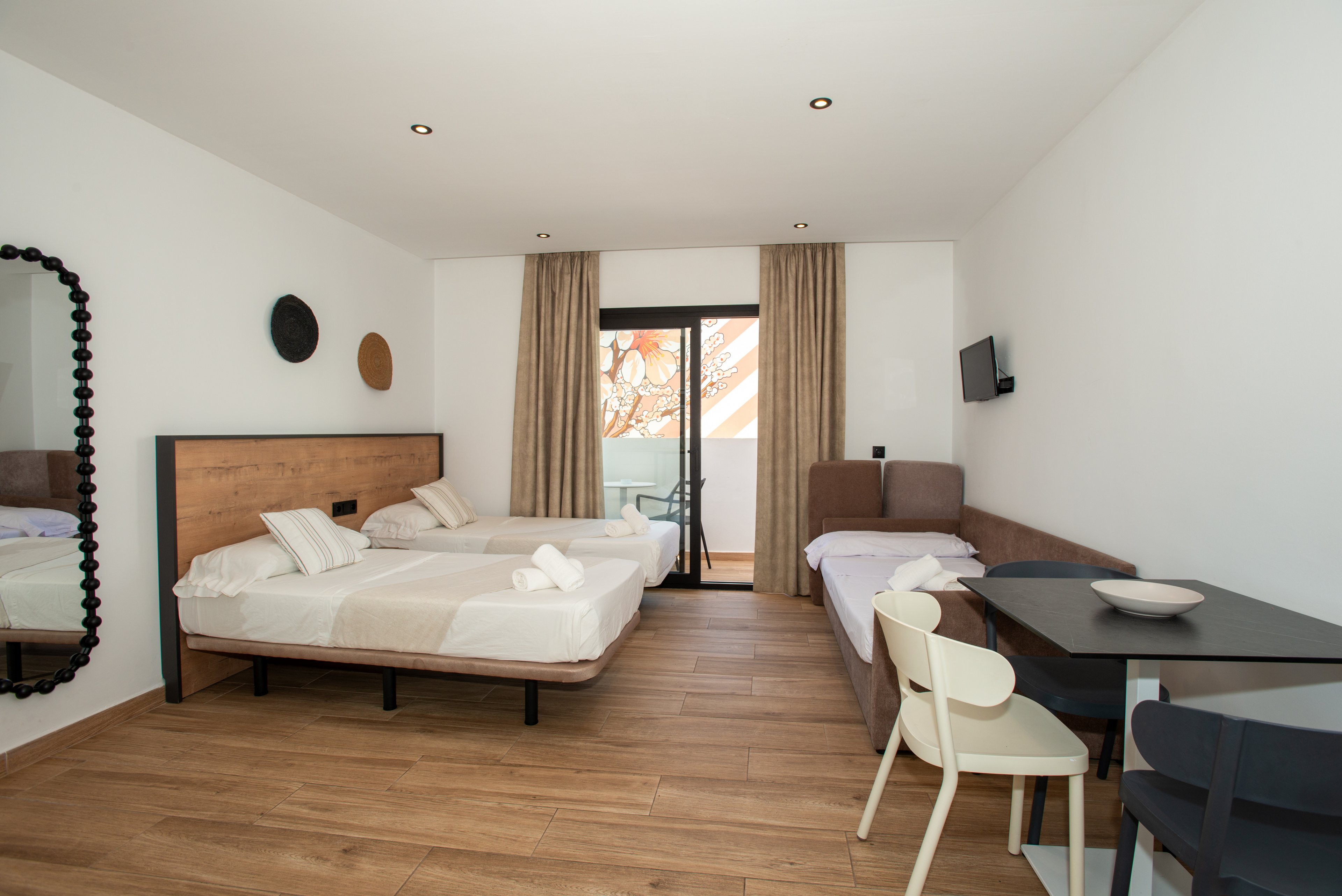 A contemporary hotel room features a double bed and a single bed with neatly folded white towels. The space has wooden flooring, simple white walls, and neutral-toned bedding for a clean, minimalist look. A small dining table with chairs and a wall-mounted television provide additional comfort. Floor-to-ceiling curtains frame a glass door leading to a balcony with an outdoor view.
