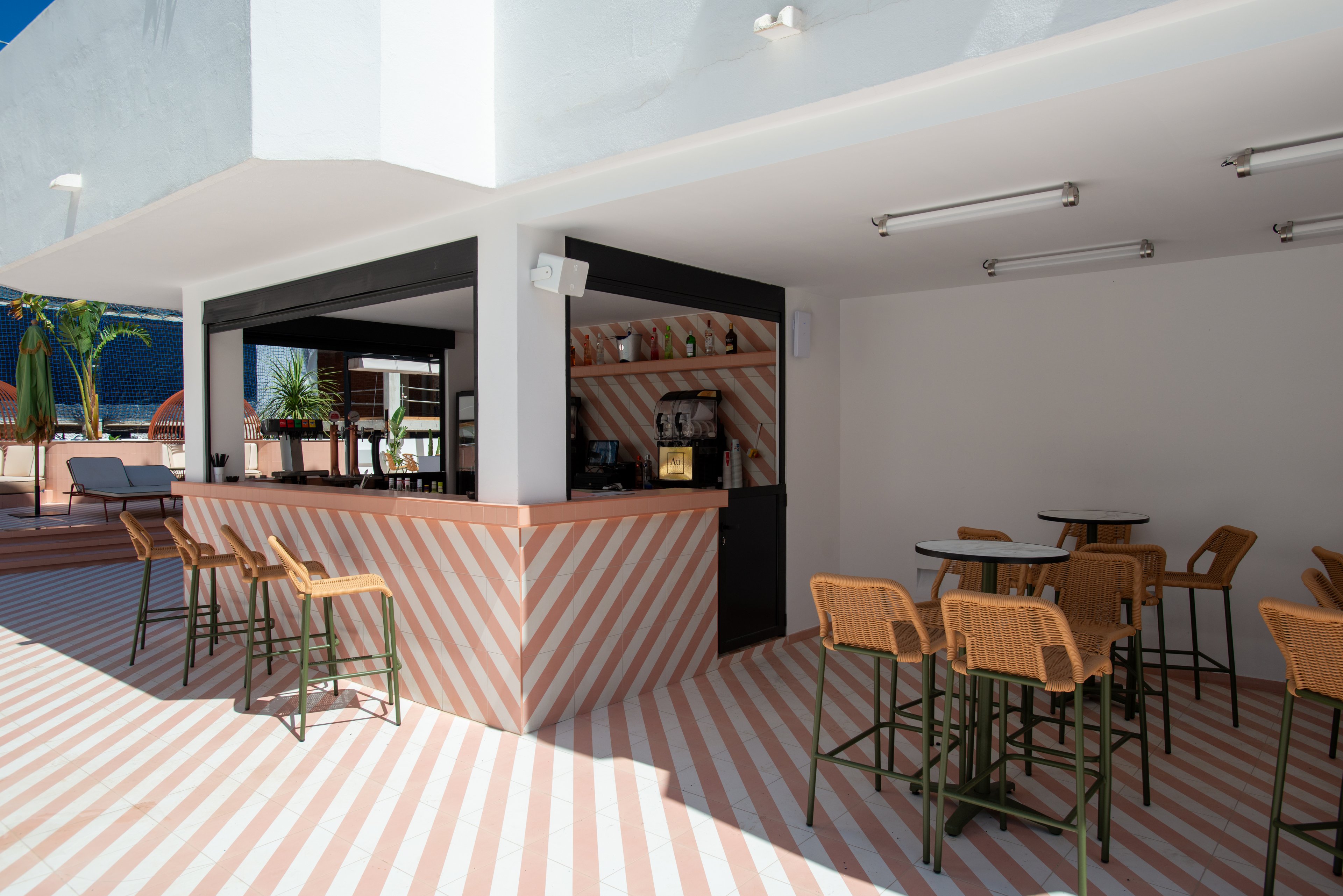 A contemporary outdoor bar stands on a terrace with striped flooring and matching bar front. High stools and small tables with woven seats are arranged around the open-air counter. The space is bright and minimal, with white walls, geometric lines, and a few potted plants adding greenery. Sunlight and clear skies create a relaxed, vacation-like atmosphere in this stylish hospitality setting.