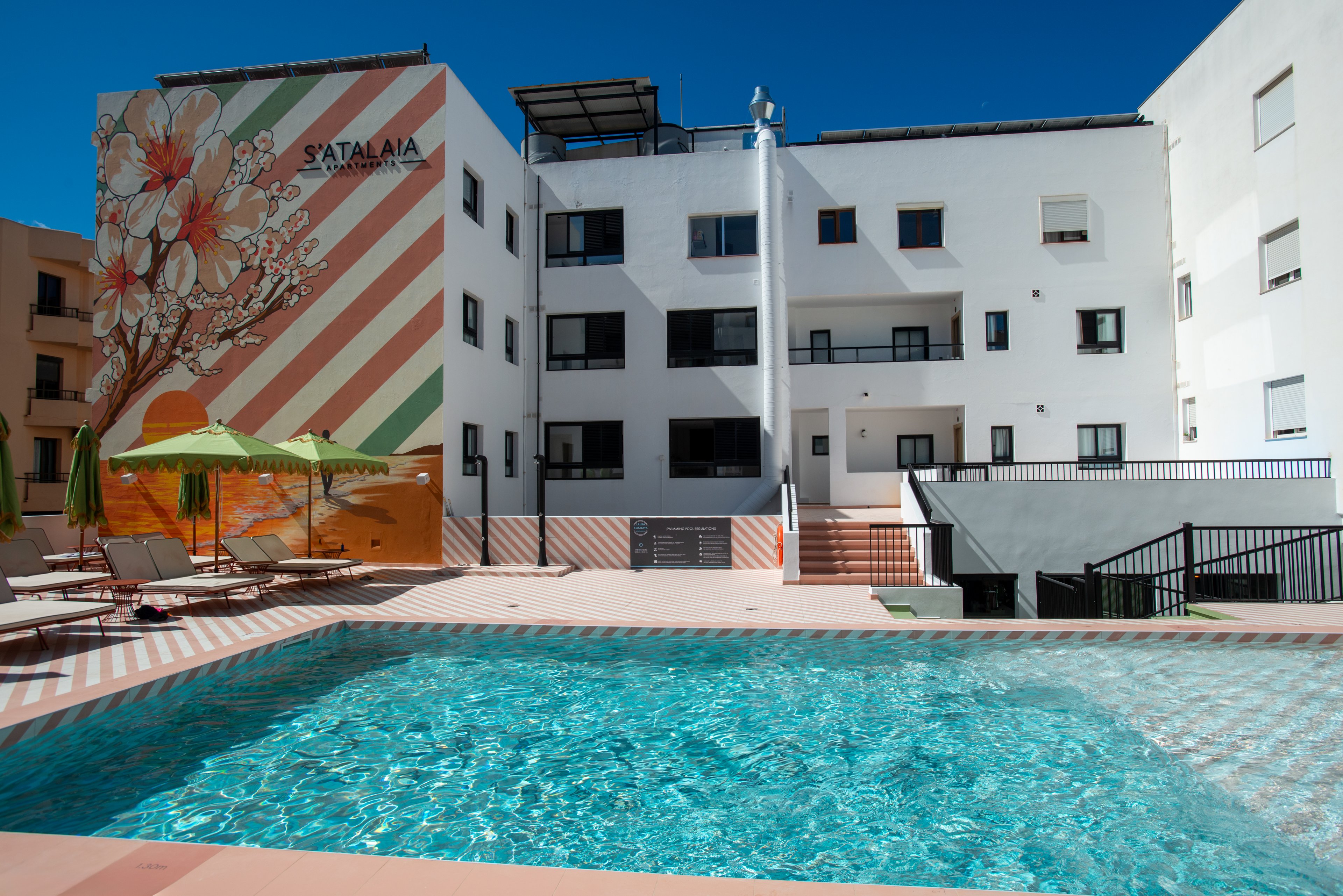 A modern white apartment building features a bright outdoor swimming pool in the foreground. The facade includes a large colorful mural with flowers and diagonal stripes, along with the visible text "SATALIA". Sun loungers and green umbrellas line the poolside terrace under a clear blue sky. The scene suggests a sunny, relaxed holiday atmosphere at a residential or vacation complex.