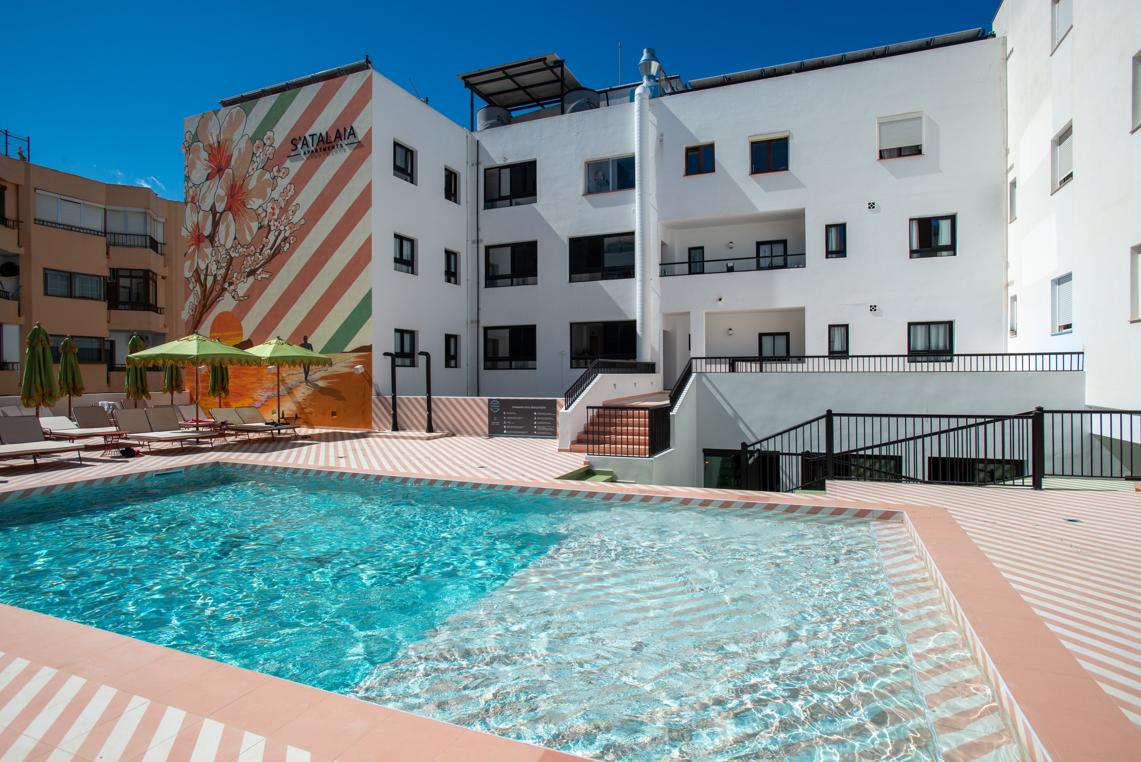 An outdoor swimming pool sits in the courtyard of a modern apartment building. The pool is surrounded by a spacious tiled deck with lounge chairs and green umbrellas. One building wall features a large colorful mural with diagonal stripes and floral artwork. Bright sunlight and a clear blue sky create a warm, inviting atmosphere for relaxation.