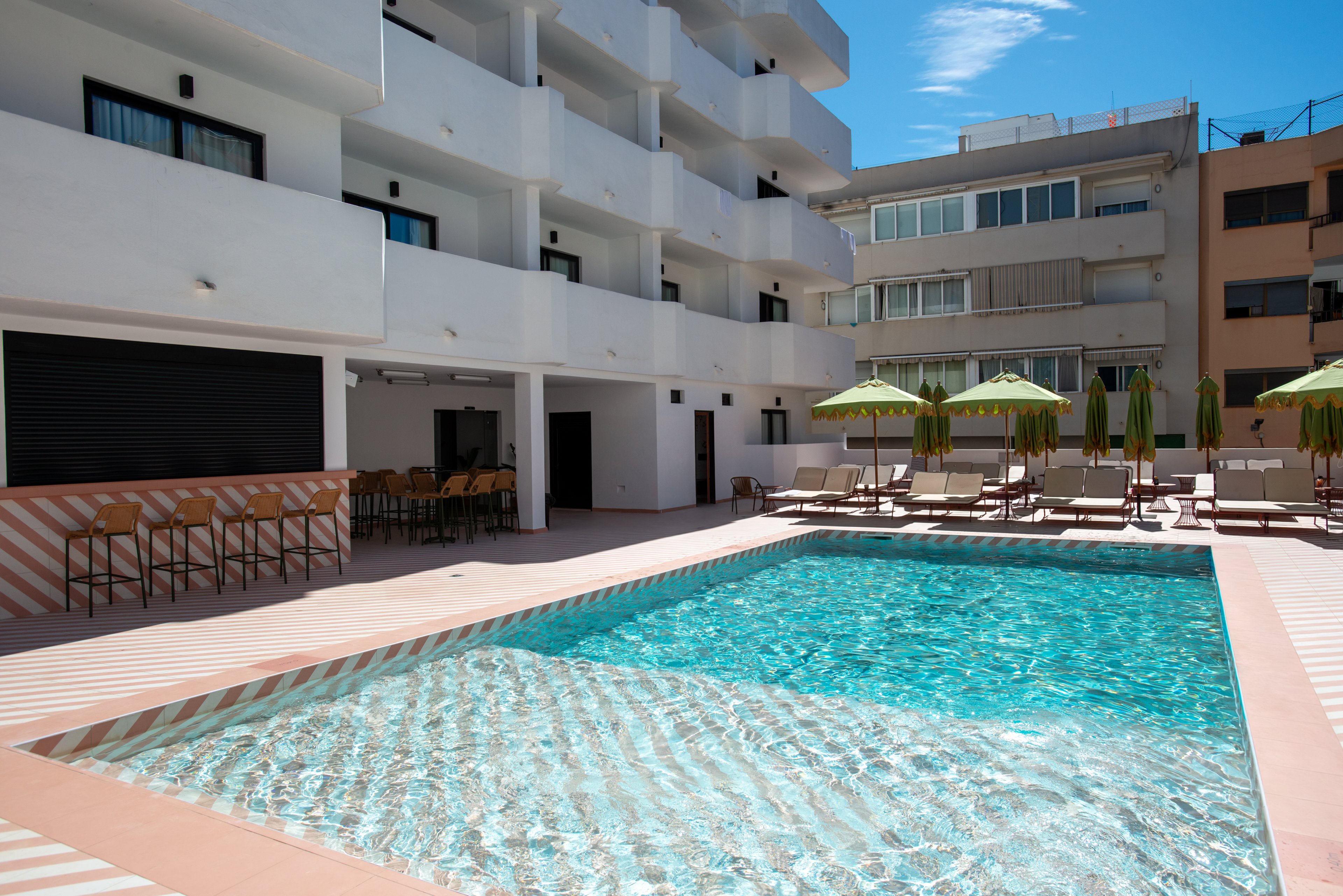 A clear blue swimming pool sits in the courtyard of a modern multi-story hotel. Sun loungers and green umbrellas line the poolside deck, suggesting a relaxed vacation setting. A small outdoor bar area with high stools is visible to the left. Surrounding buildings and bright daylight create a warm, summery atmosphere ideal for leisure and relaxation.