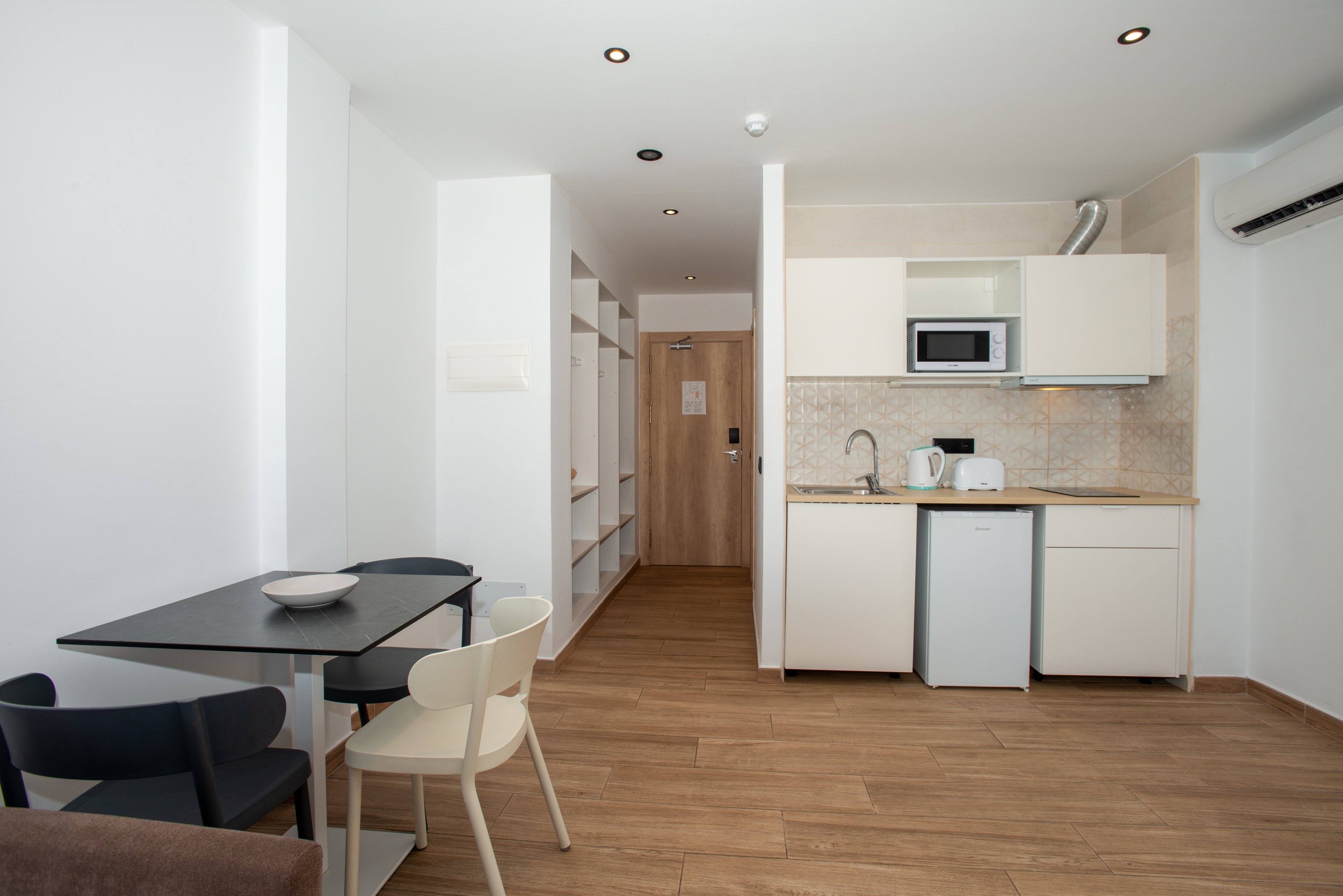 Compact modern studio interior featuring a small kitchenette and dining table. The kitchen area includes white cabinets, a sink, microwave, kettle, and a mini fridge beneath a tiled backsplash. A simple black dining table with mismatched chairs sits on warm wood-look flooring. Recessed ceiling lights and a wall-mounted air conditioner brighten and cool the minimalist space, with open shelving and a wooden door visible in the background.