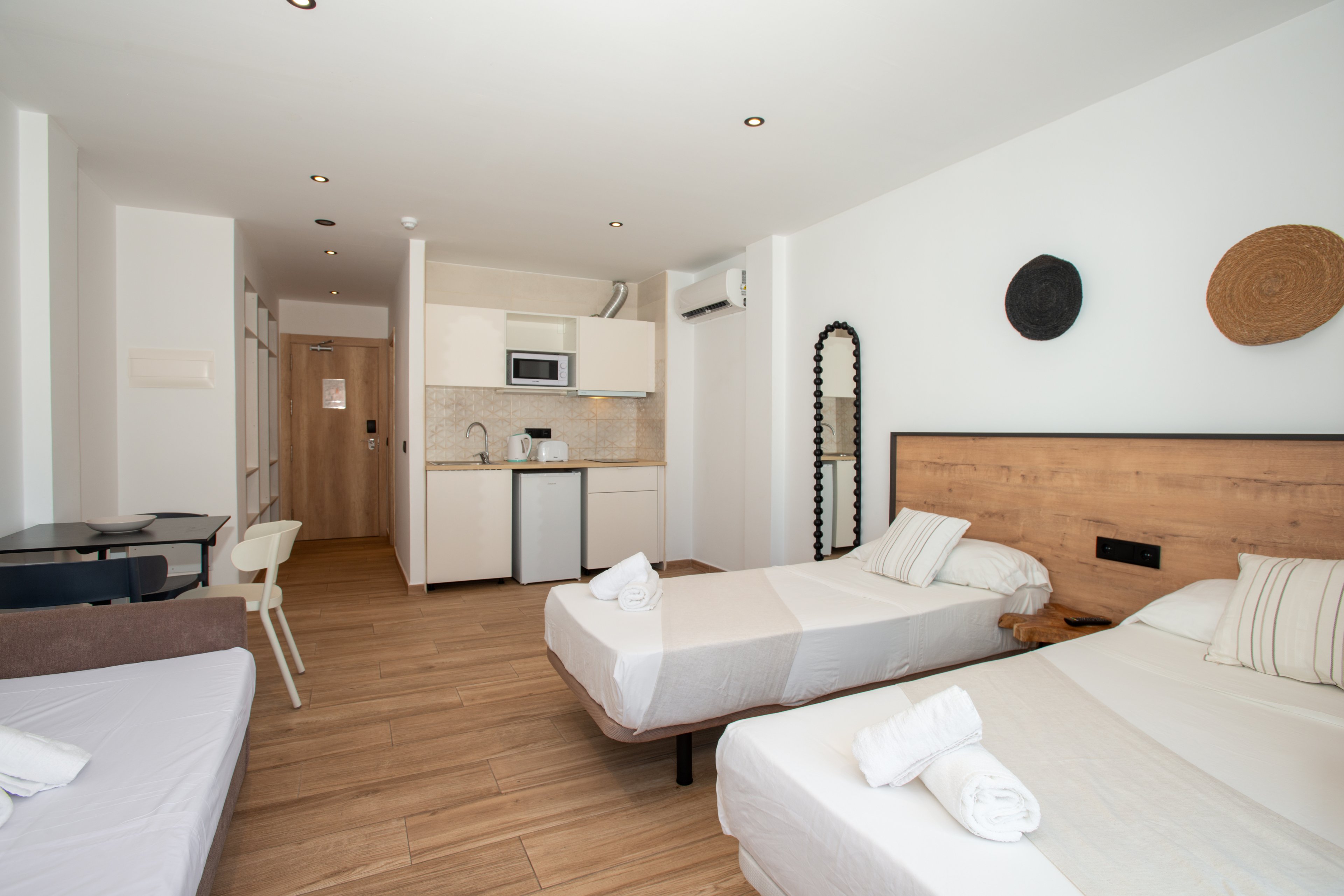 Spacious studio-style hotel room with two single beds and a sofa bed is shown in a bright, minimalist design. The space includes a small kitchenette with a sink, microwave, and mini fridge near the entrance door. Light wood flooring and white walls create an airy, clean atmosphere, complemented by simple wall decor and recessed ceiling lights. Rolled white towels on the beds and neutral textiles emphasize comfort and readiness for guests.