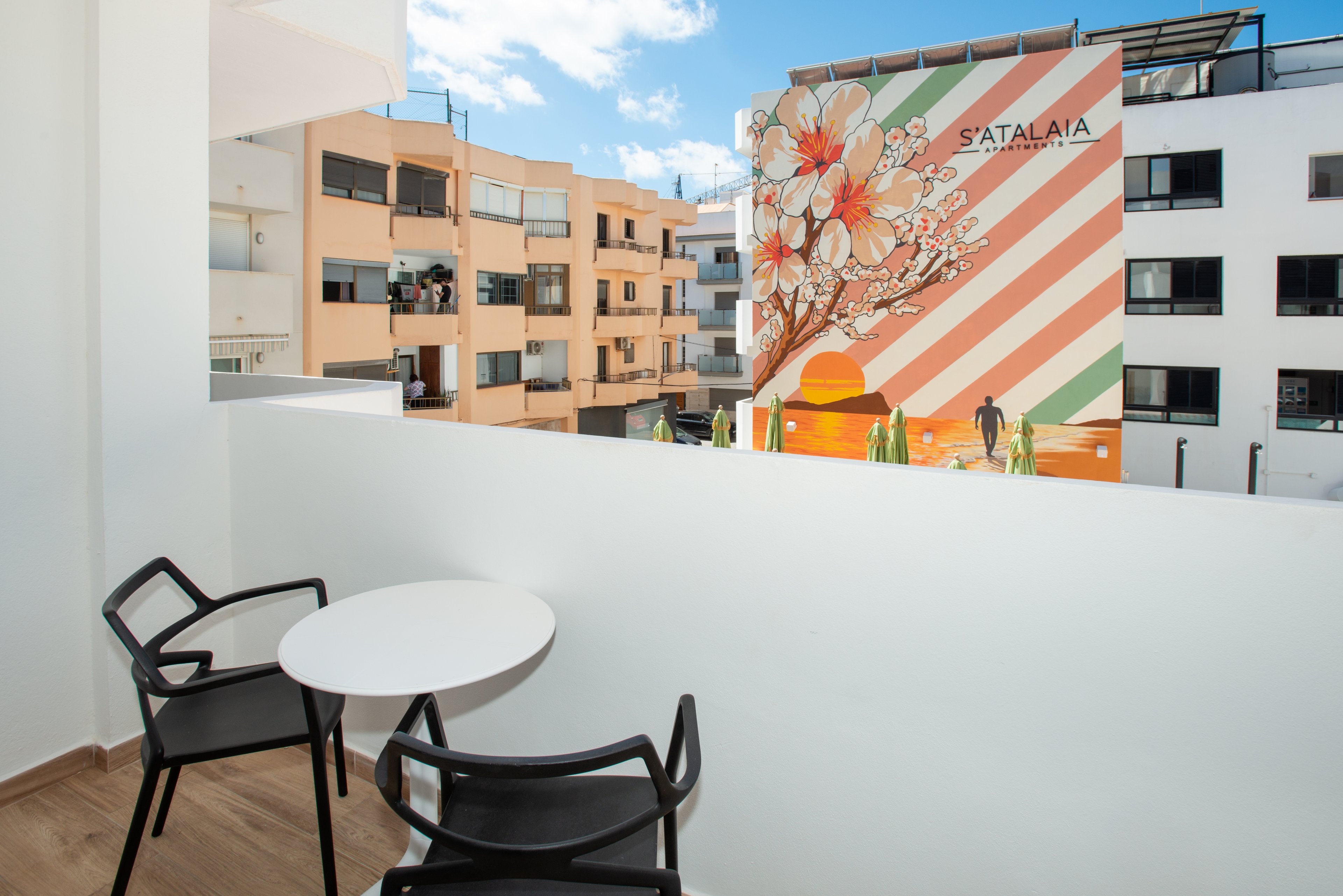 A small modern balcony features a round white table and two black chairs overlooking a residential courtyard. Across from the balcony, a large building facade displays a colorful floral mural with diagonal stripes. The word "SATALAIA" is clearly visible on the mural. Bright daylight and a blue sky with scattered clouds create a cheerful urban atmosphere. The scene emphasizes contemporary apartment living with artistic outdoor views.