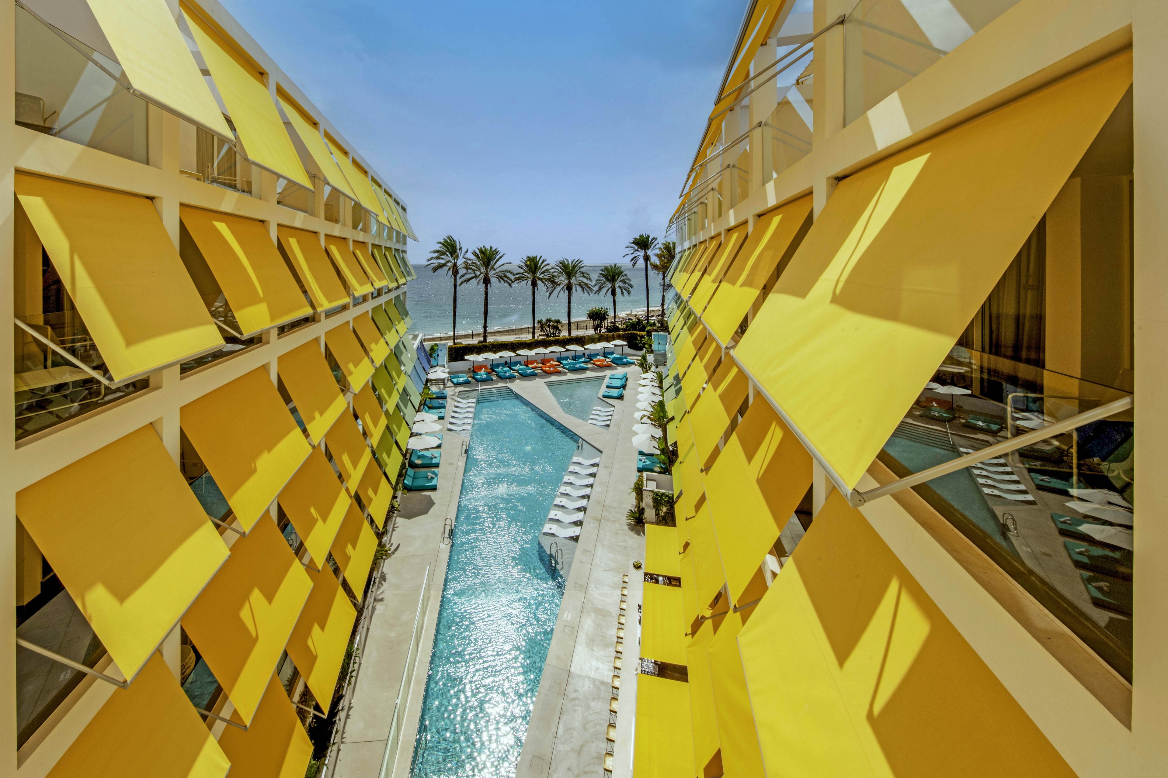 A contemporary hotel courtyard features a long central swimming pool framed by two buildings with large yellow sunshades. Lounge chairs and colorful poolside seating line the water, creating a vibrant resort atmosphere. Tall palm trees stand at the far end of the pool, leading the eye toward the distant ocean and clear blue sky. The strong geometric lines and bold yellow and turquoise palette give the scene a modern, architectural feel.