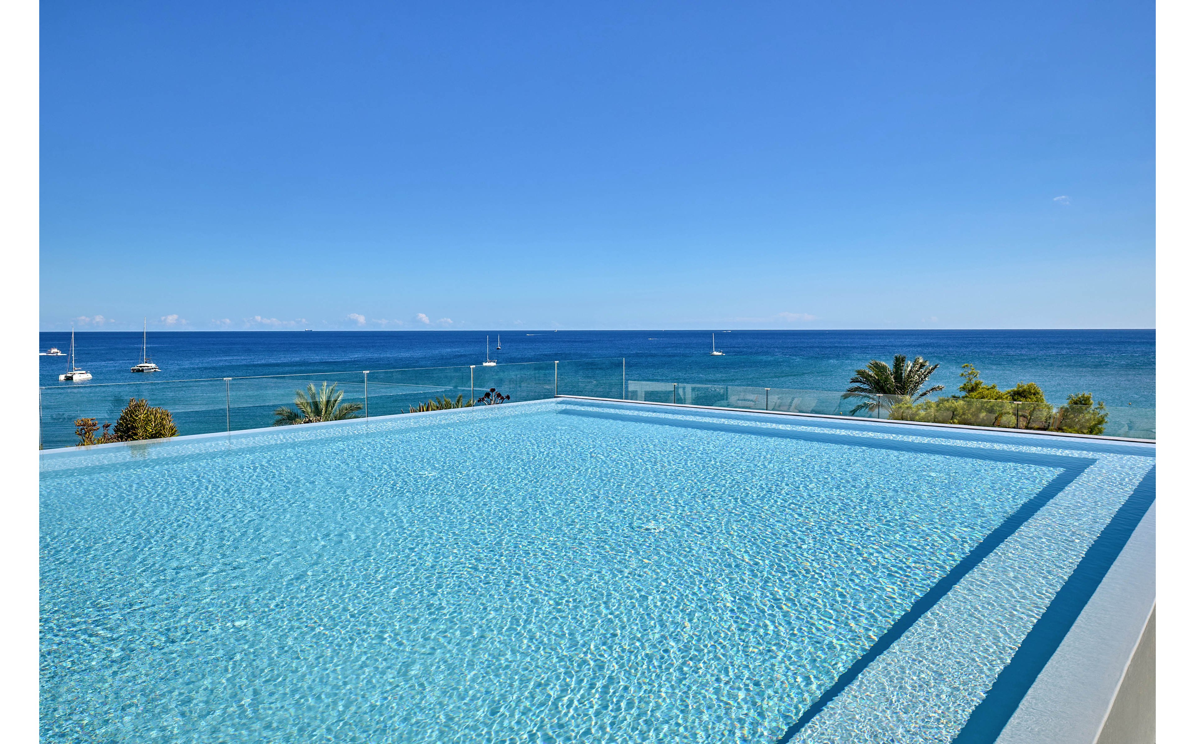 A tranquil infinity pool stretches toward a clear blue ocean under a cloudless sky. The water in the pool is still and sparkling, seamlessly blending with the distant horizon. Sailboats and small vessels are visible on the sea, adding subtle activity to the serene coastal scene. Palm trees and shoreline vegetation frame the edge of the view, enhancing the tropical resort atmosphere.
