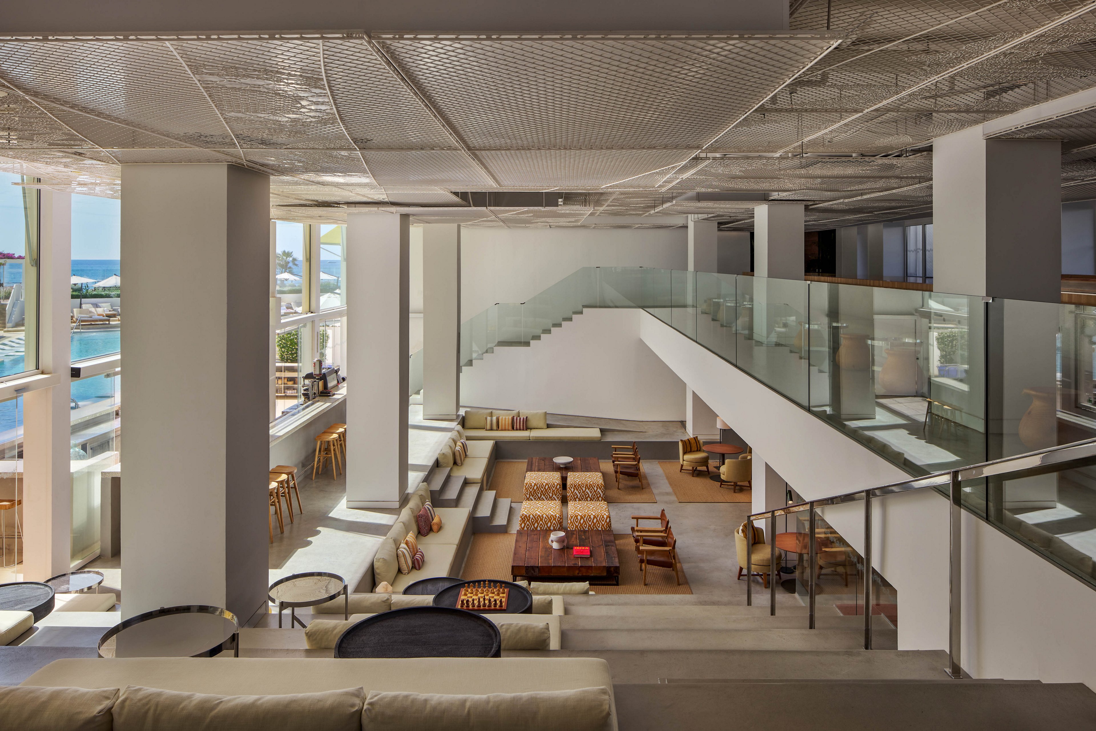 Spacious contemporary lounge area features multiple seating levels and a central staircase with glass railings. Large windows flood the interior with natural light and offer views of the ocean and nearby marina. Neutral tones, wood accents, and minimalist furniture create a clean, modern aesthetic. The space appears calm and inviting, suitable for relaxing, socializing, or informal meetings.