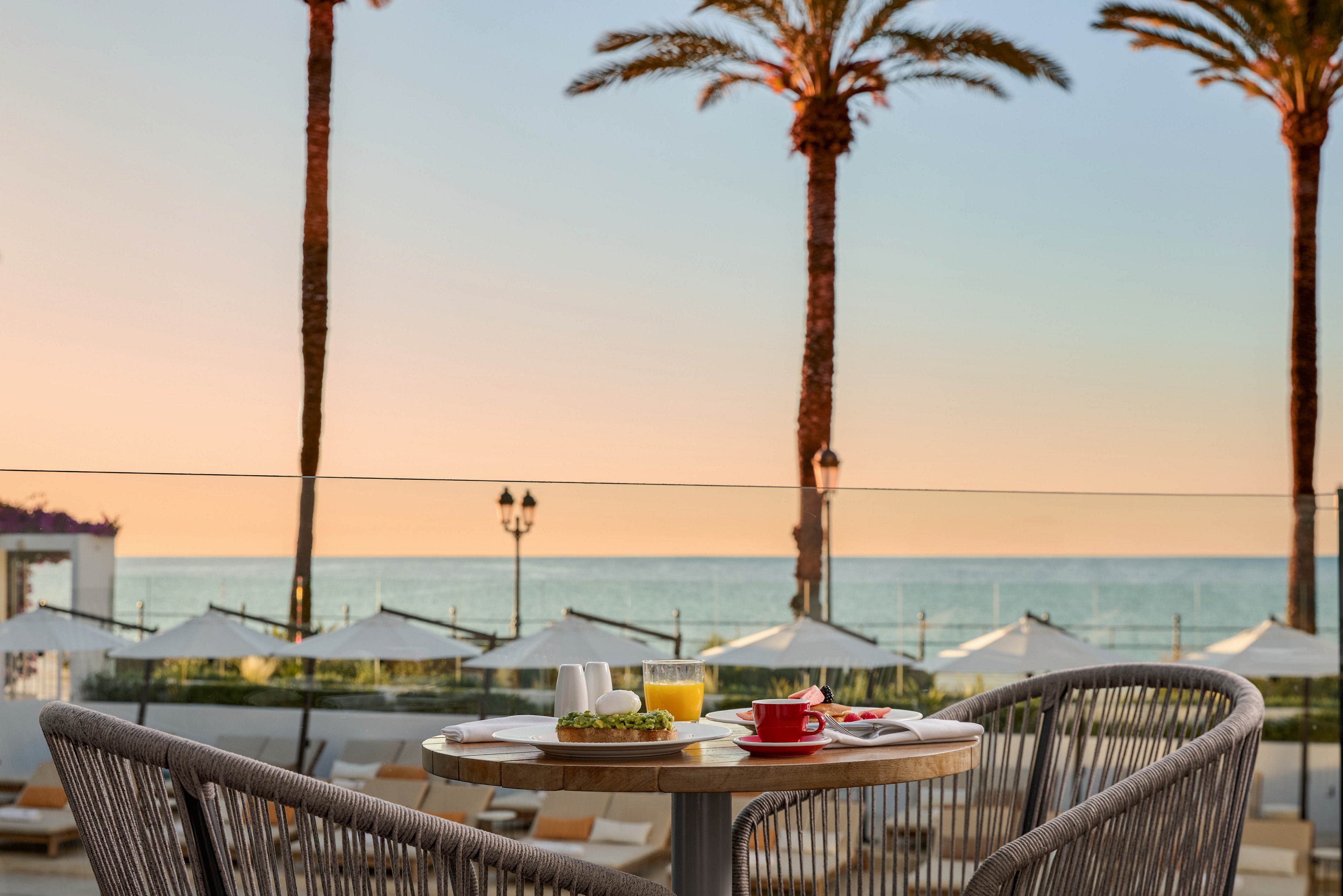 A small round table is set with breakfast items, including a red coffee cup, juice, and a plate of food. The table sits on a terrace with woven chairs facing the ocean. Tall palm trees and rows of white beach umbrellas line the shore in the background. The sky glows with soft pastel colors, creating a calm and relaxing seaside atmosphere.