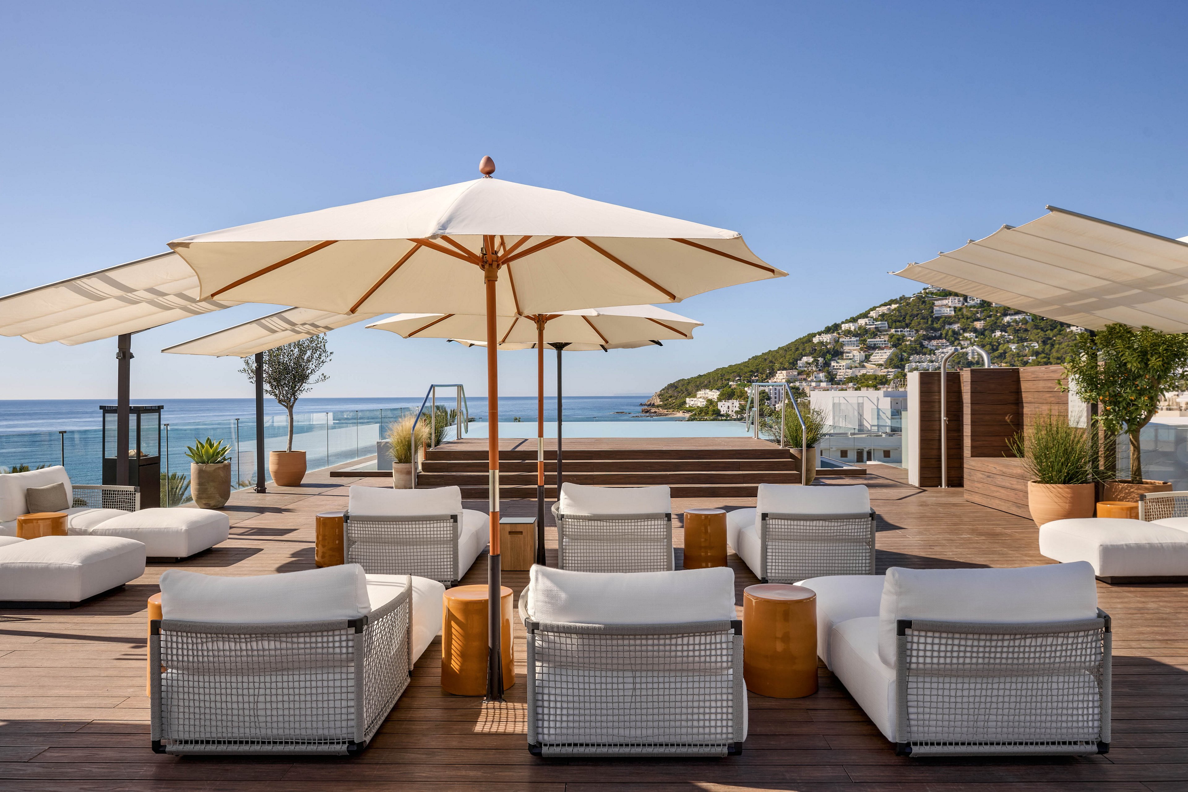 Modern white lounge chairs and umbrellas line a spacious rooftop terrace with a view of the ocean. A sleek infinity pool stretches toward the horizon, framed by wooden decking and minimalist planters. The scene is bright and sunny, with clear blue skies and calm water creating a serene atmosphere. Hillside buildings in the background suggest an upscale coastal resort destination.