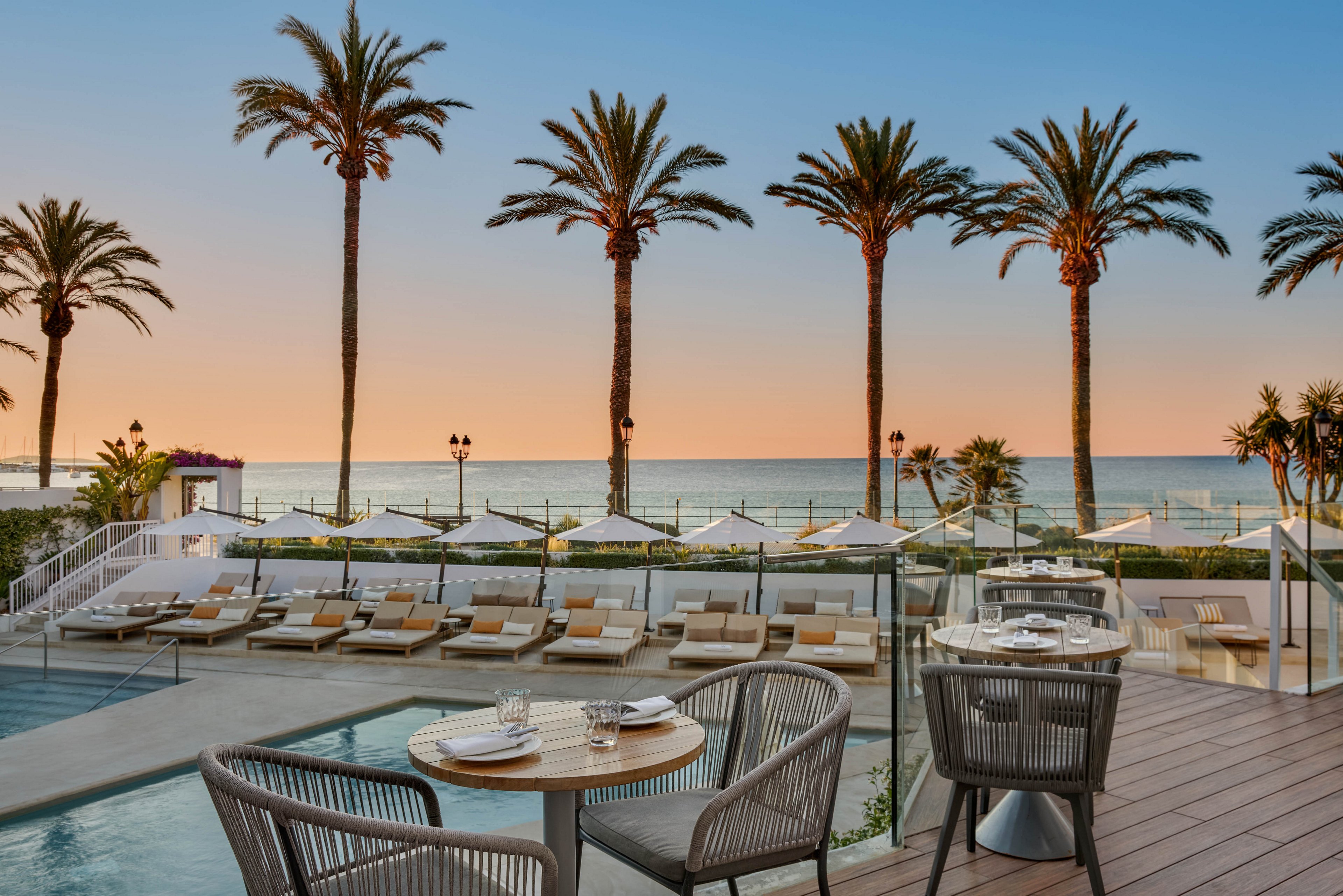 An elegant seaside resort terrace overlooks a calm ocean at sunset. A small pool and neatly arranged sun loungers with umbrellas create a relaxing beachfront atmosphere. In the foreground, a set dining table with modern chairs suggests an upscale outdoor restaurant experience. Tall palm trees frame the scene against a pastel sky, emphasizing a tranquil vacation mood.
